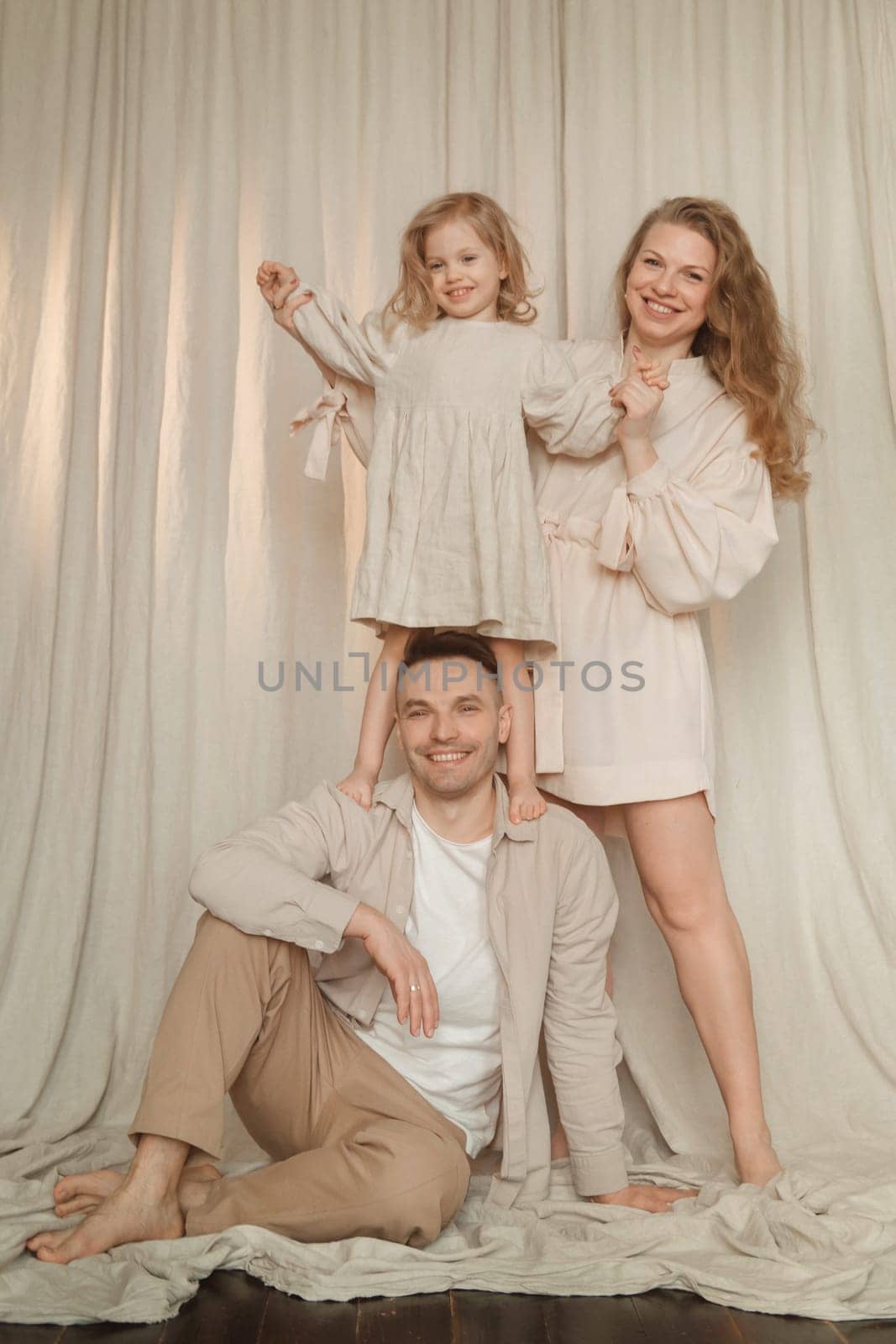 A family of three enjoys a joyful moment indoors, with a child sitting on a parent's shoulders while the other parent stands beside them. The setting features soft drapes and a relaxed atmosphere.