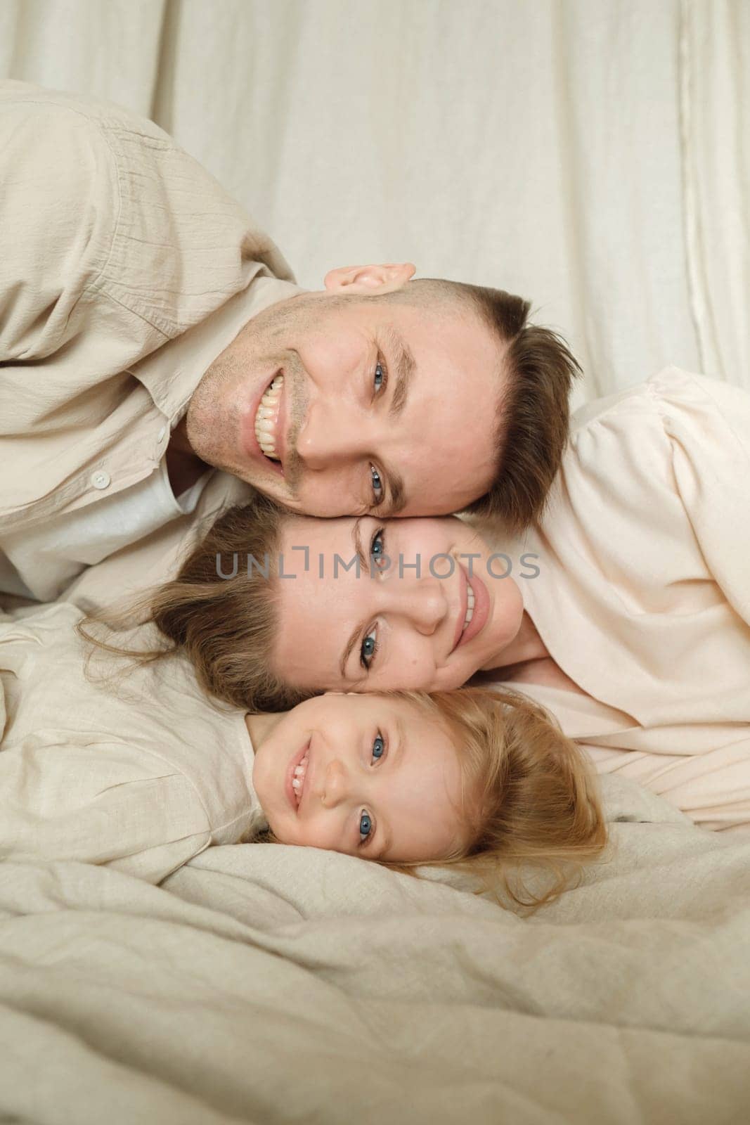 A joyful family of three enjoys a playful moment together, lying on a soft blanket indoors. Their beaming smiles and close proximity showcase a strong bond filled with love.