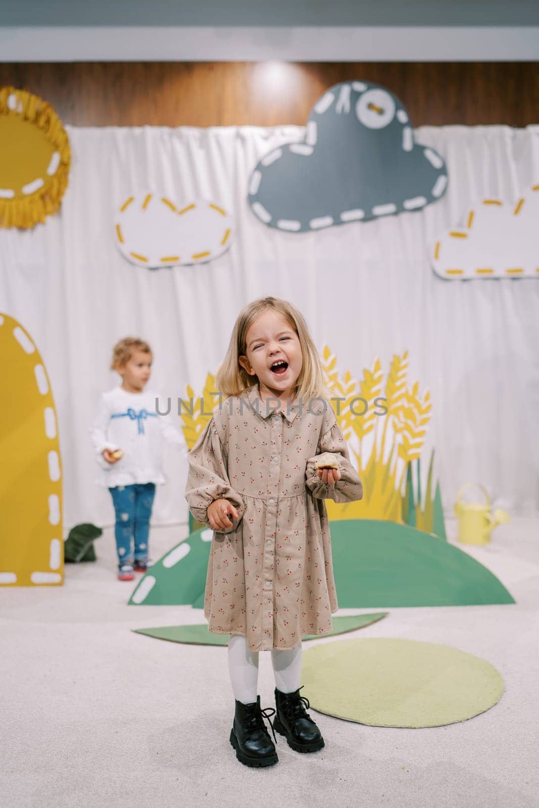 Little laughing girl with a bun in her hand stands on stage among colorful decorations in a kindergarten. High quality photo