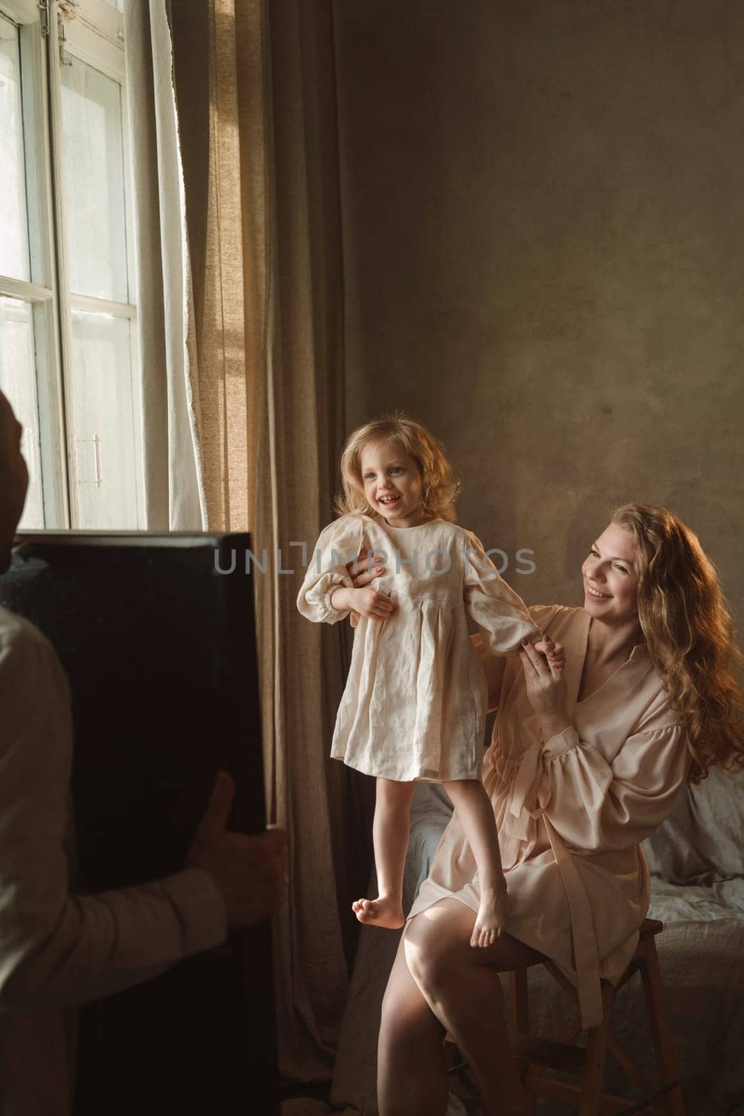 In a bright room, a mother joyfully lifts her daughter as they share a playful moment. The father captures their happiness, creating lasting memories in a loving family setting.