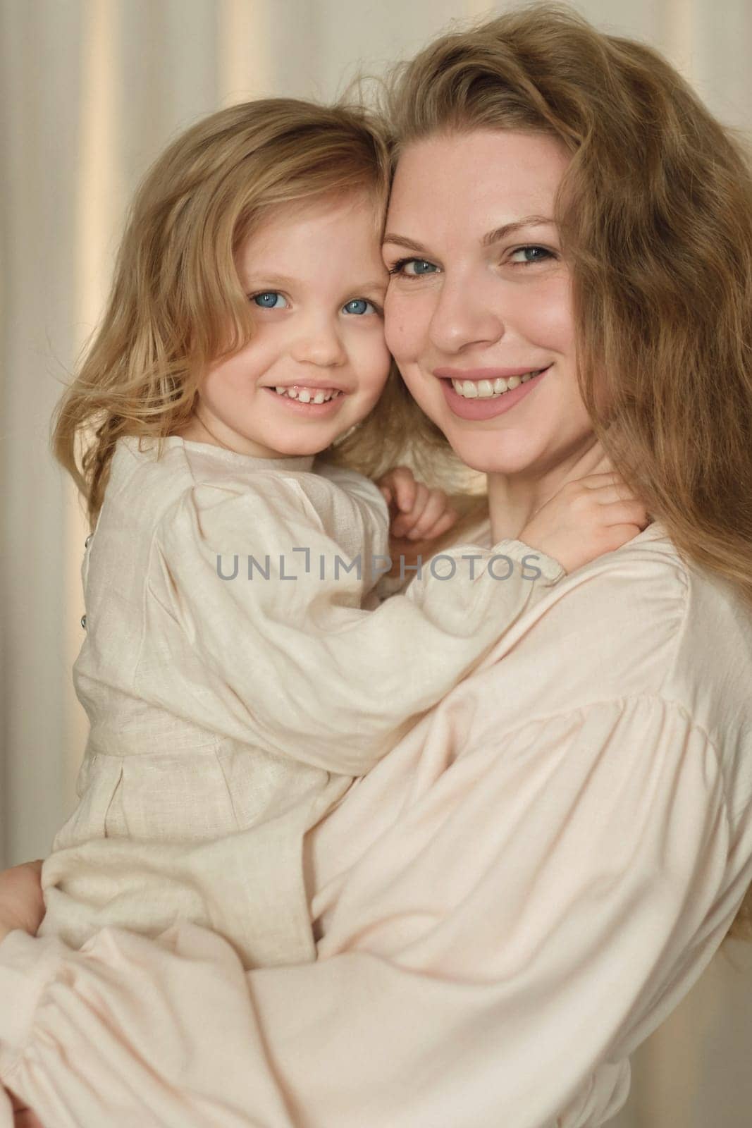 A mother holds her young daughter in a warm embrace, both smiling brightly. The soft lighting creates an intimate atmosphere, showcasing their close connection in a home setting.