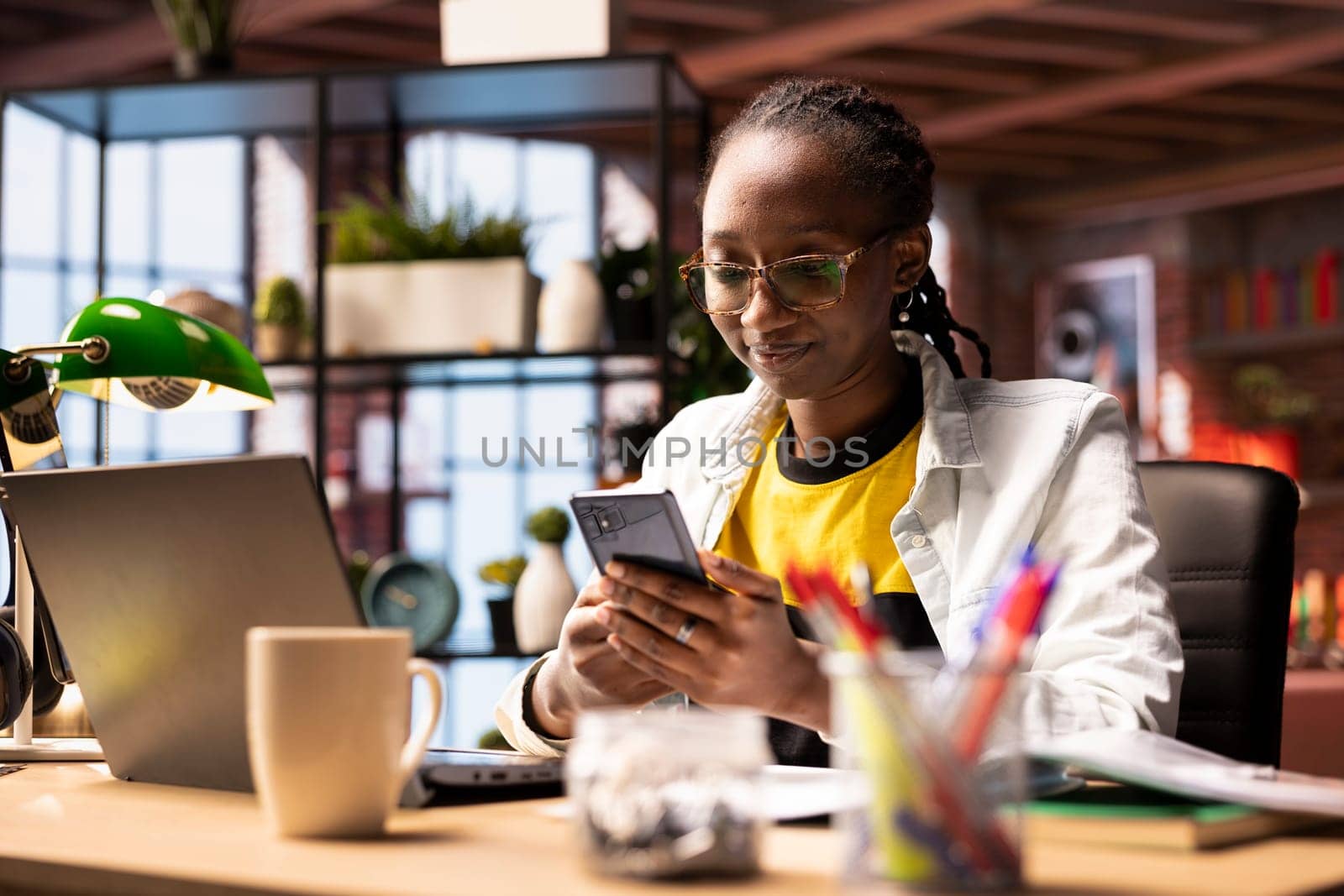 Smiling woman in home office using smartphone to compose social media posts, producing content. African american person monitoring social media engagement using mobile phone