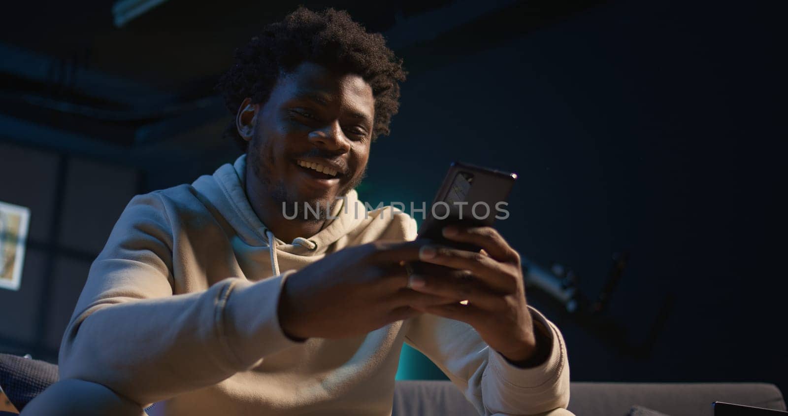 Relaxed black freelancer chatting with friends on his smartphone via texts, sitting on the couch. Young adult showcasing laughter and leisure, staying entertained while working remotely. Camera B.