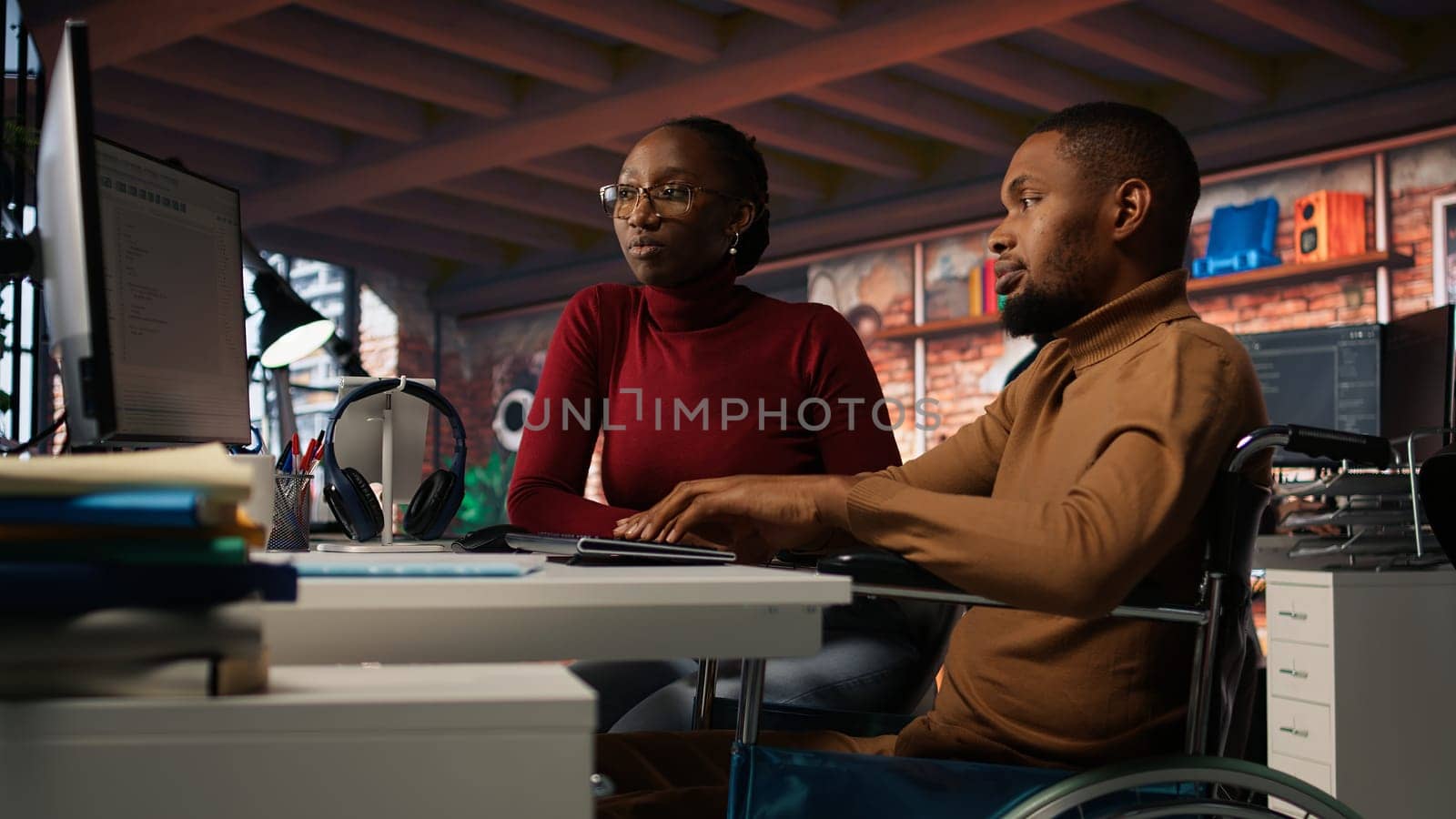 Manager coming to inclusive startup office employee in wheelchair desk to offer help. Thoughtful supervisor checking up on software development agency worker with disability, camera A