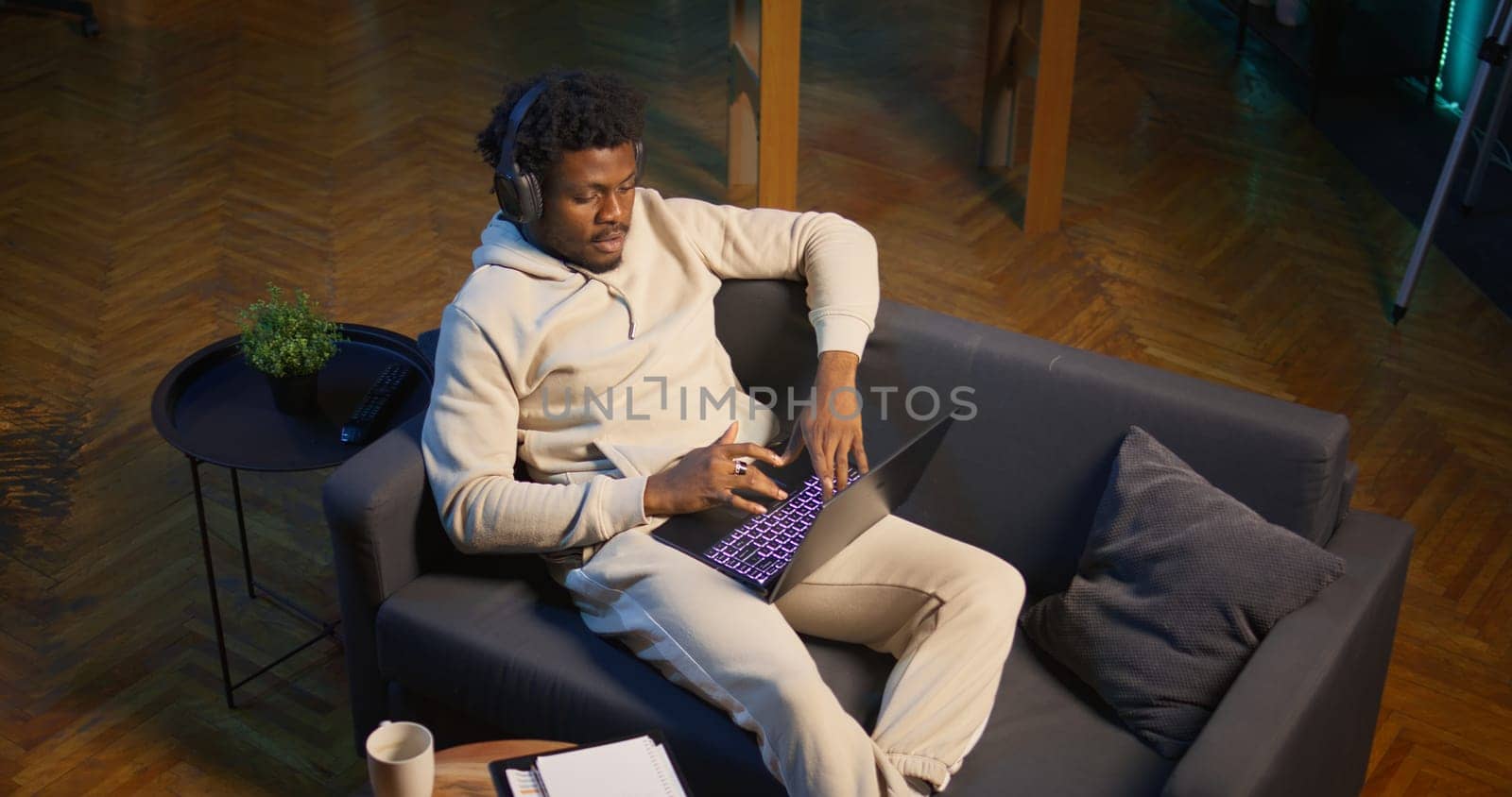 African american self employed man browses the web and listens to music, having fun with songs on headset at his modern home. Young adult laying on couch and relaxing in the living room. Camera B.