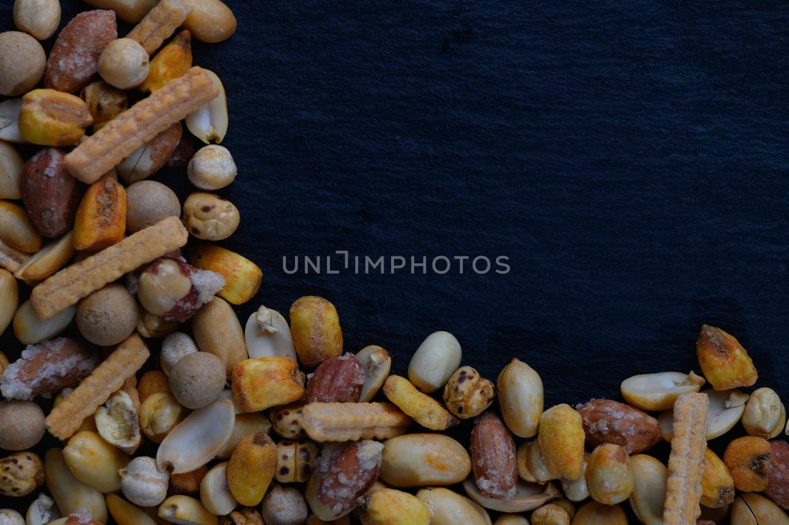 A vibrant mix of nuts and crispy snacks is artfully scattered along the edges of a dark slate surface, inviting sharing and enjoyment during lively conversations.