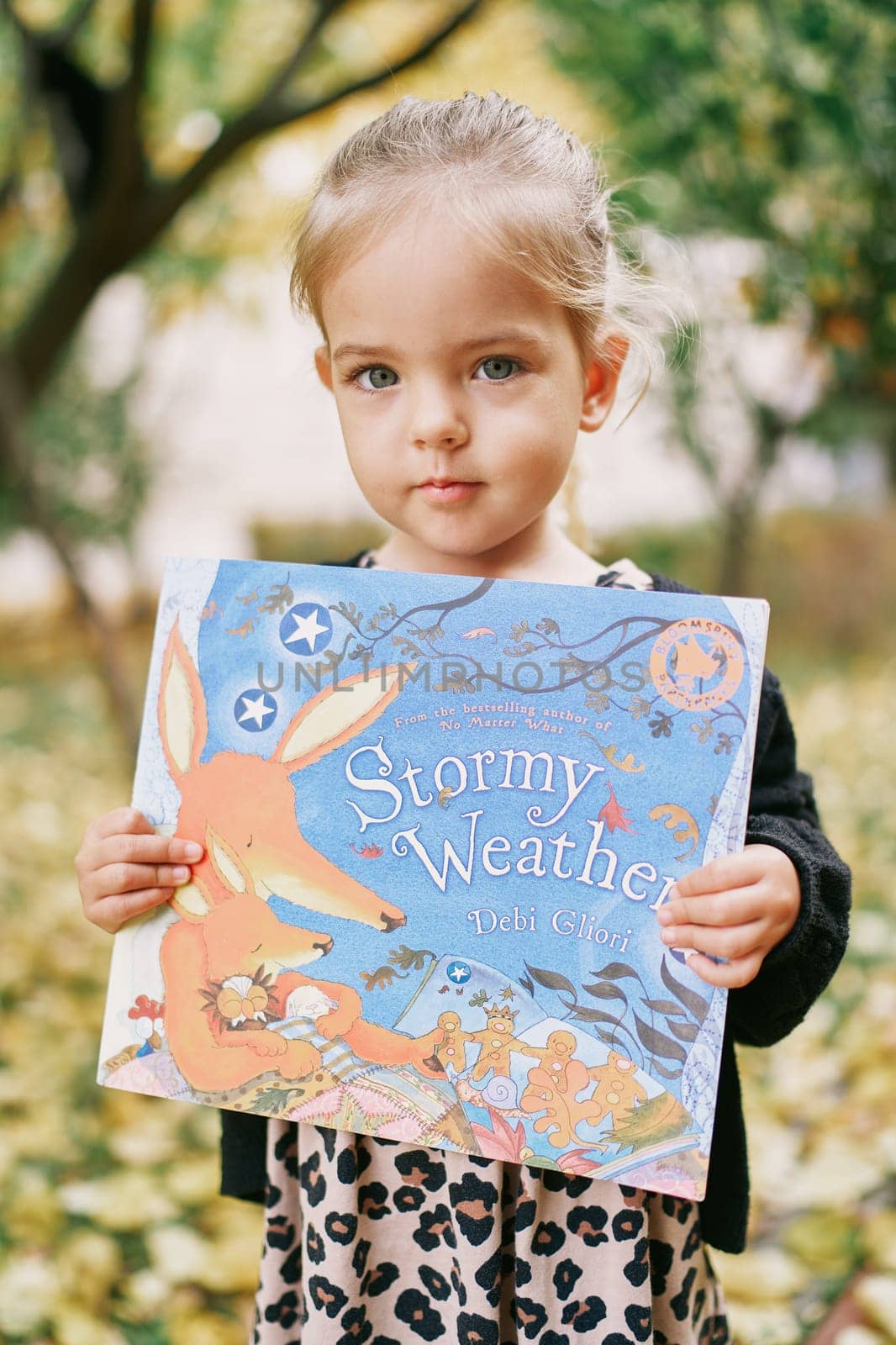 Little girl with a colorful book in her hands stands in the garden. Title: Stormy weather. Debi Gliori. High quality photo