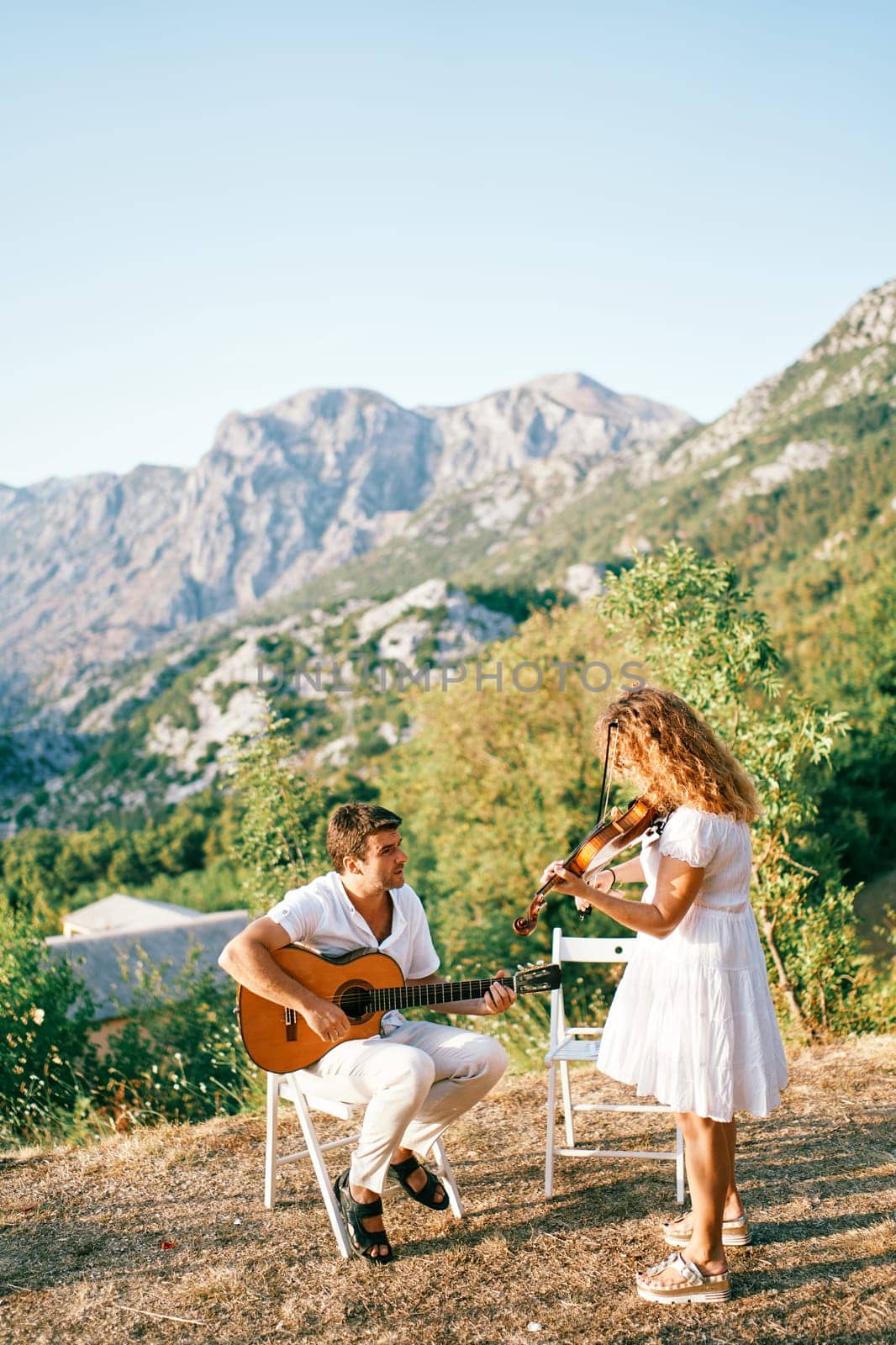 Violinist plays the violin while standing next to a seated guitarist playing the guitar in the mountains. High quality photo