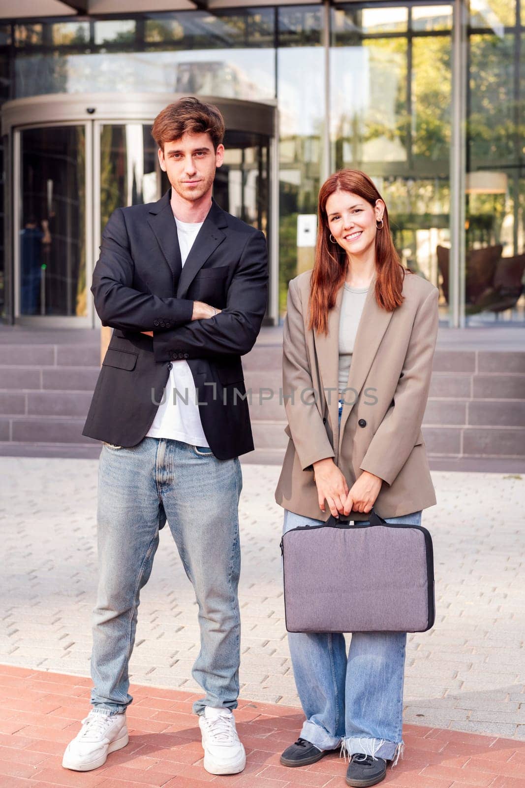 business man and woman standing outside of an office building smiling happy while looking at camera, they are wearing casual clothes and she has a briefcase