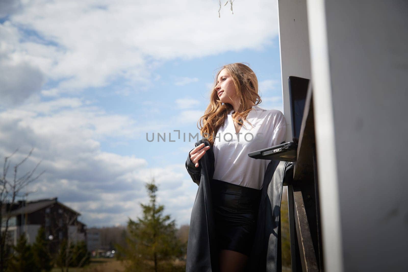 Woman stands outside on a sunny day, casually holding a laptop while enjoying the pleasant weather.