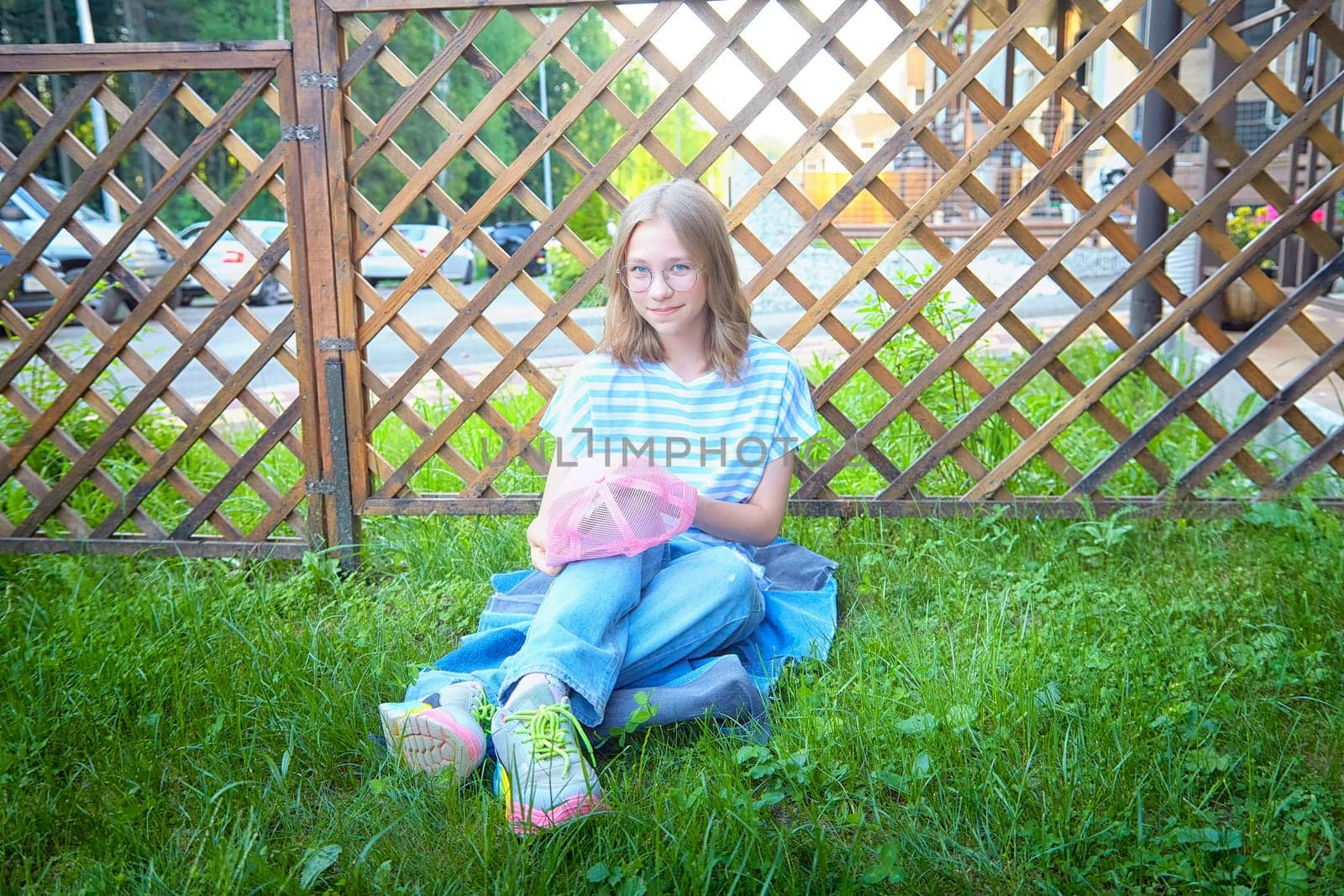 A girl sits on green grass in a casual outfit, holding a pink bag, enjoying a warm day next to a wooden lattice fence.