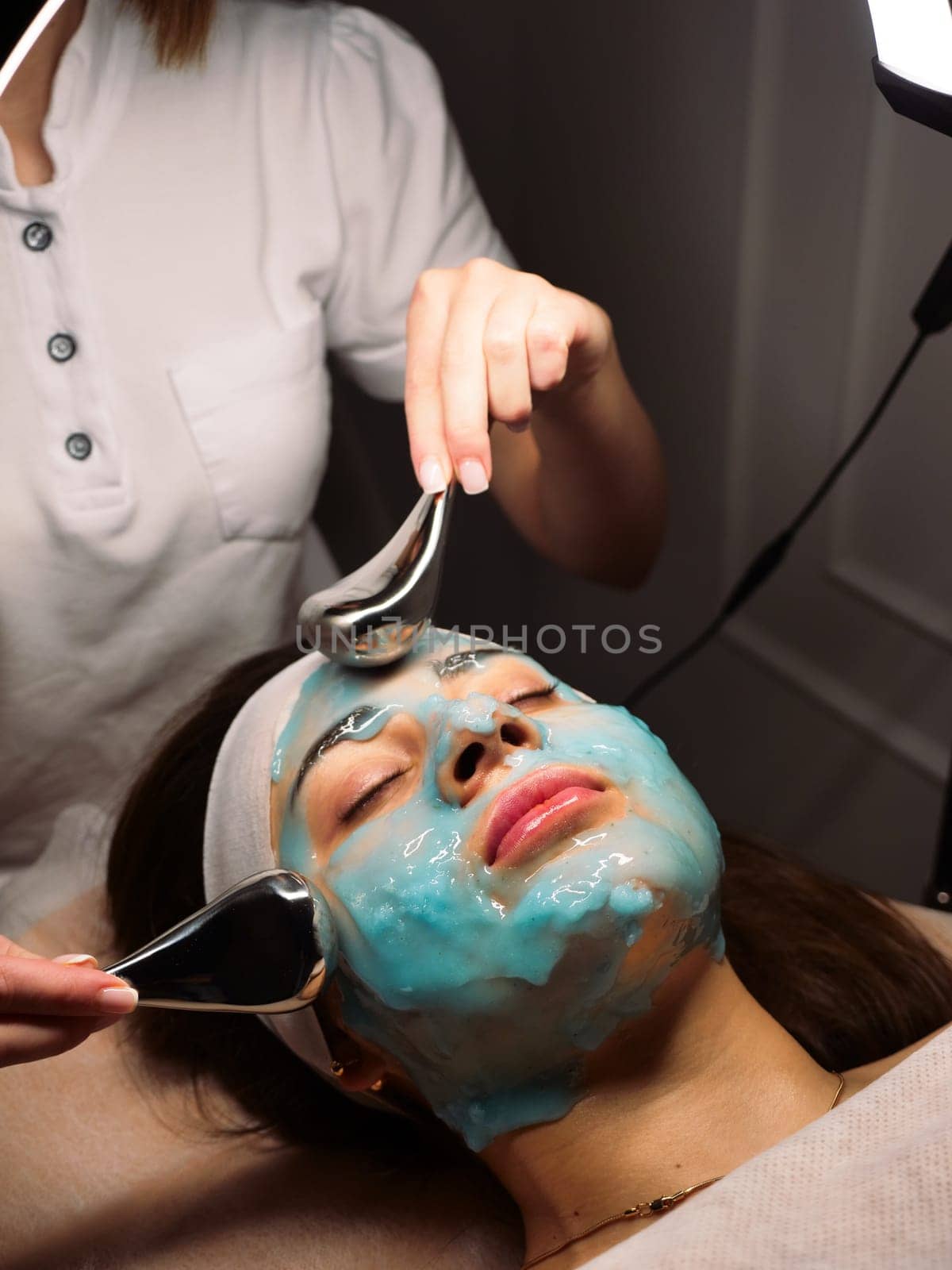Cosmetologist does cryo massage with globules on a blue hydrogel alginate mask in the beauty salon, vertically, close-up. Woman Facial Skin and Body Care