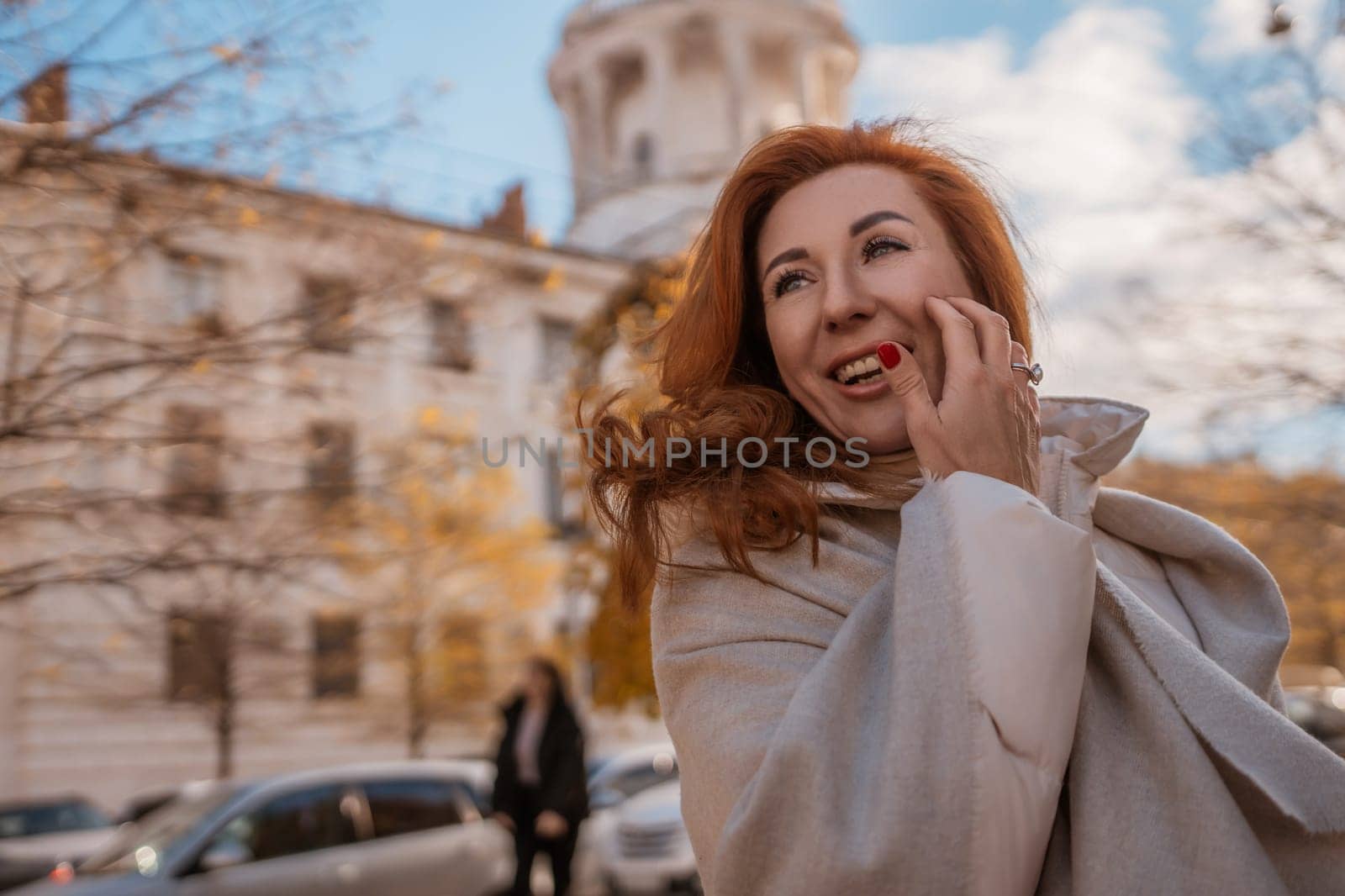 Redhead woman in a beige coat smiles, hand on face, outdoors. Old European architecture and parked cars blur in the background. Autumnal setting.