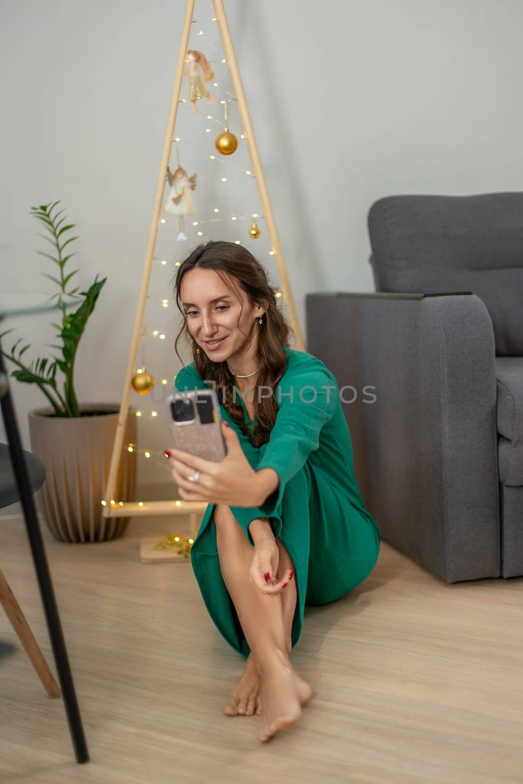 Woman in green dress sits barefoot on floor, smiling at phone, beside a minimalist gold Christmas tree.