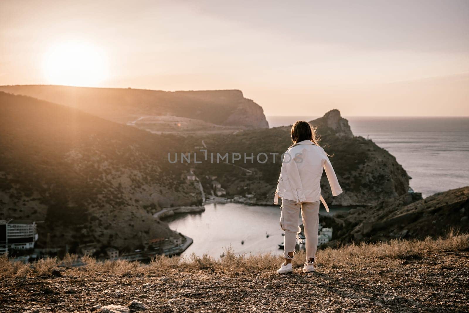 A woman stands on a hill overlooking a body of water. The sun is setting in the background, casting a warm glow over the scene. The woman is enjoying the view and taking in the beauty of the landscape