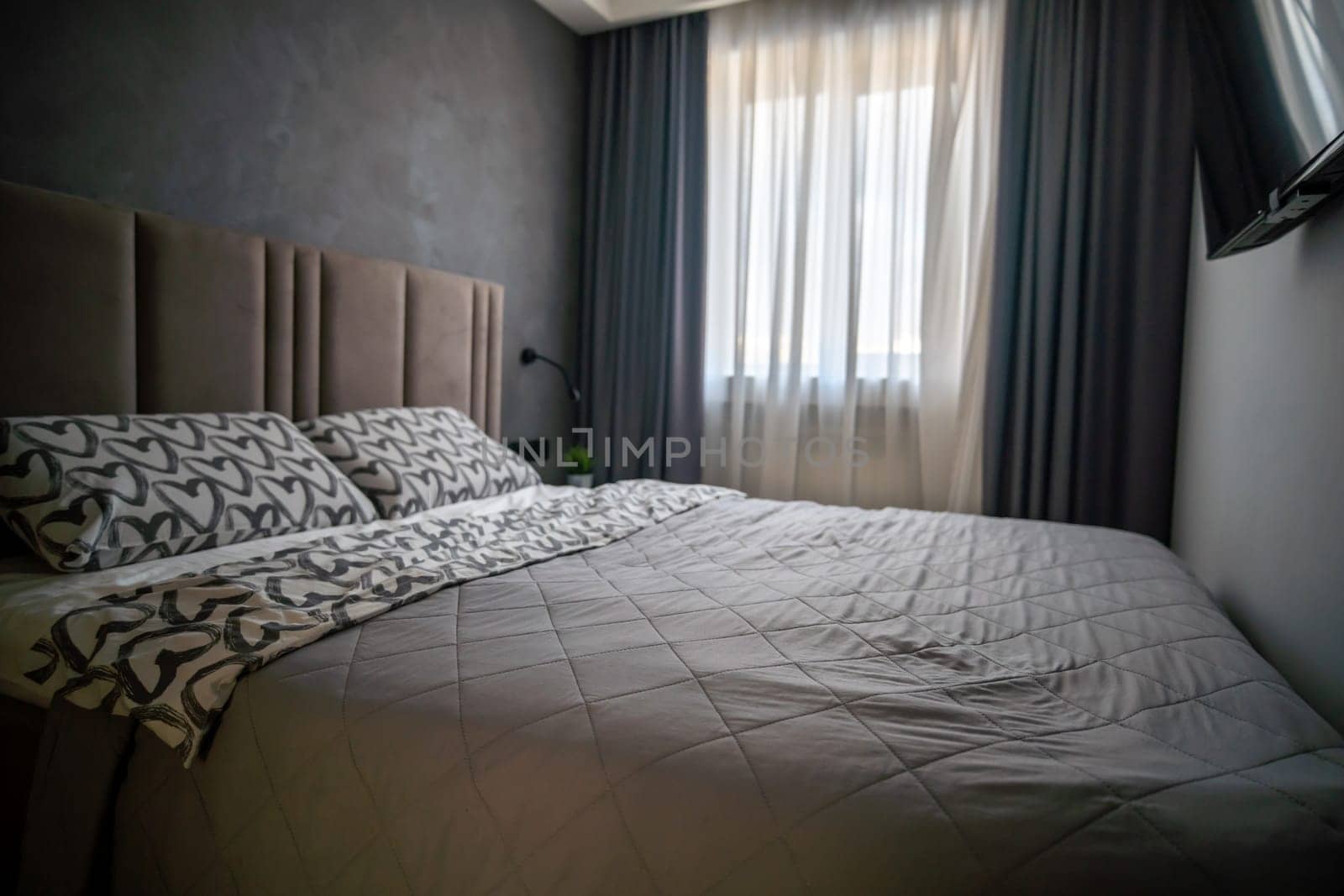 Modern bedroom with a double bed, patterned bedding, textured headboard, dark walls, sheer curtains, and a glimpse of a television.