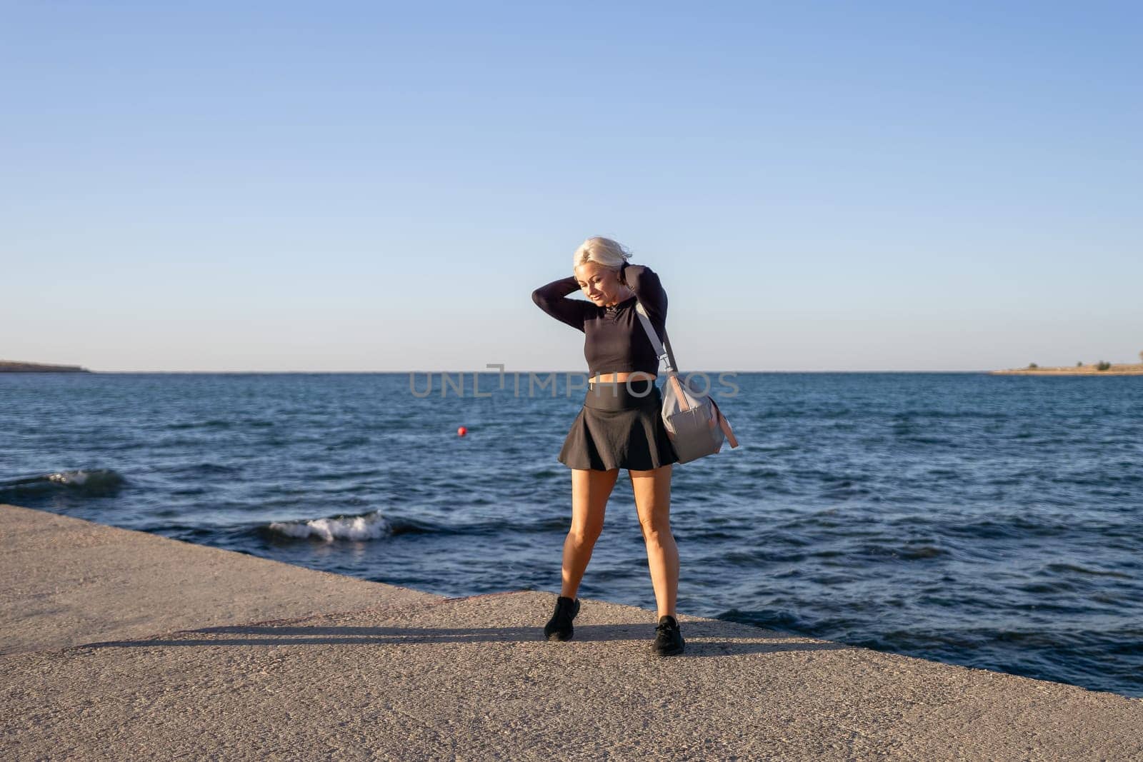 A blonde woman in a black crop top and skirt stands on a concrete pier overlooking a blue ocean. She has a bag slung over her shoulder and is holding her hair. The sky is blue and there are waves crashing on the shore.