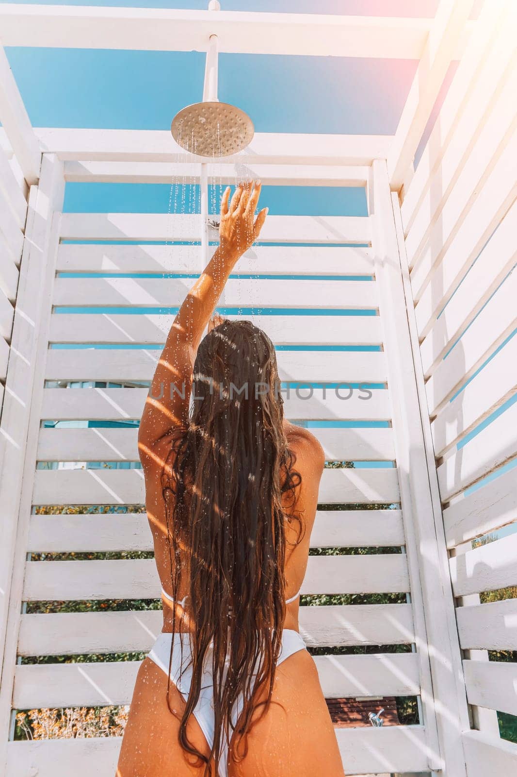 Woman with long brown hair rinsing off under an outdoor shower in a white beachside stall. Sunlight streams through the slats.