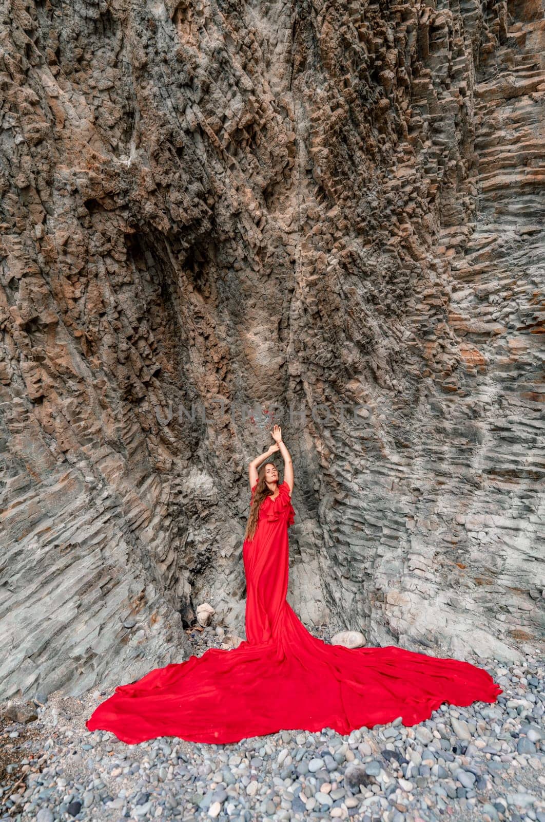 woman sea red dress. Woman with long hair on a sunny seashore in a red flowing dress, back view, silk fabric waving in the wind. Against the backdrop of the blue sky and mountains on the seashore