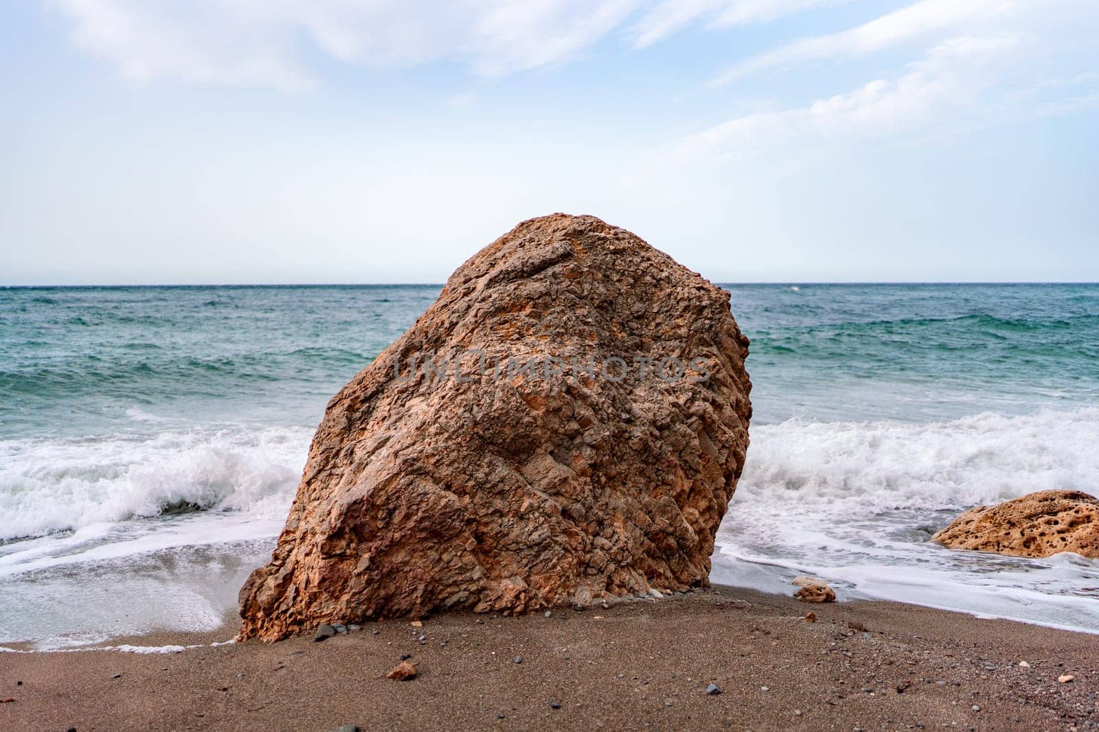 A large rock sits on the sand, with waves crashing around it at the beach against a cloudy sky.