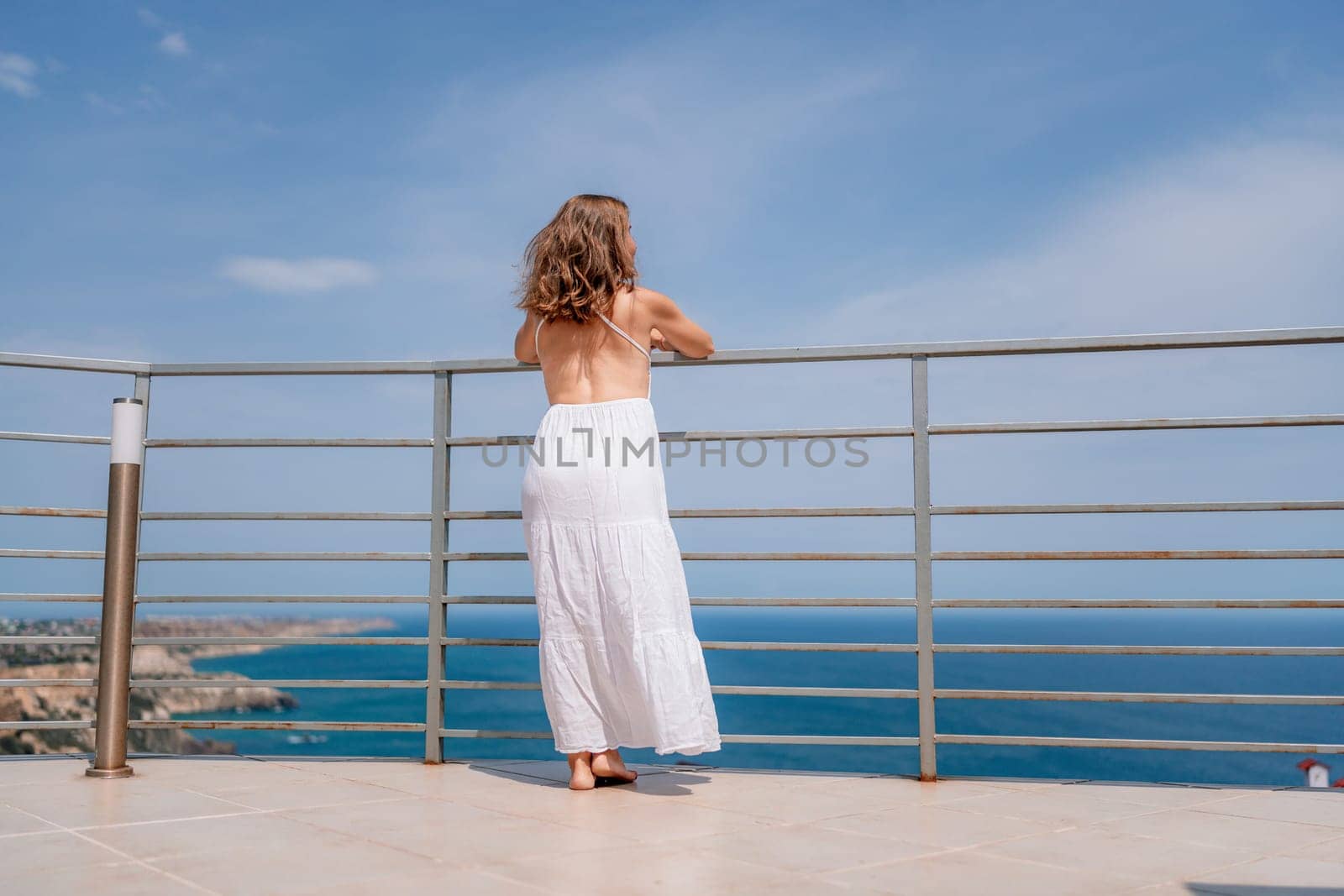 A woman in a white dress overlooks the blue ocean from a balcony with metal railings on a sunny day. The view stretches across the water.