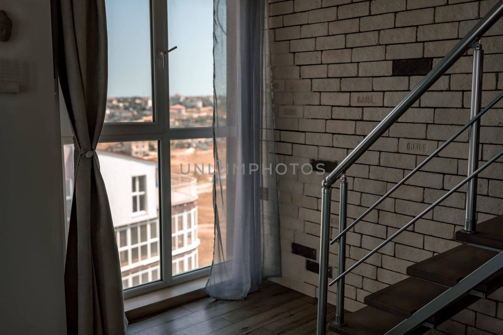A sunlit room features a window with a city view, sheer curtains, and a staircase with a metal railing against a white brick wall.