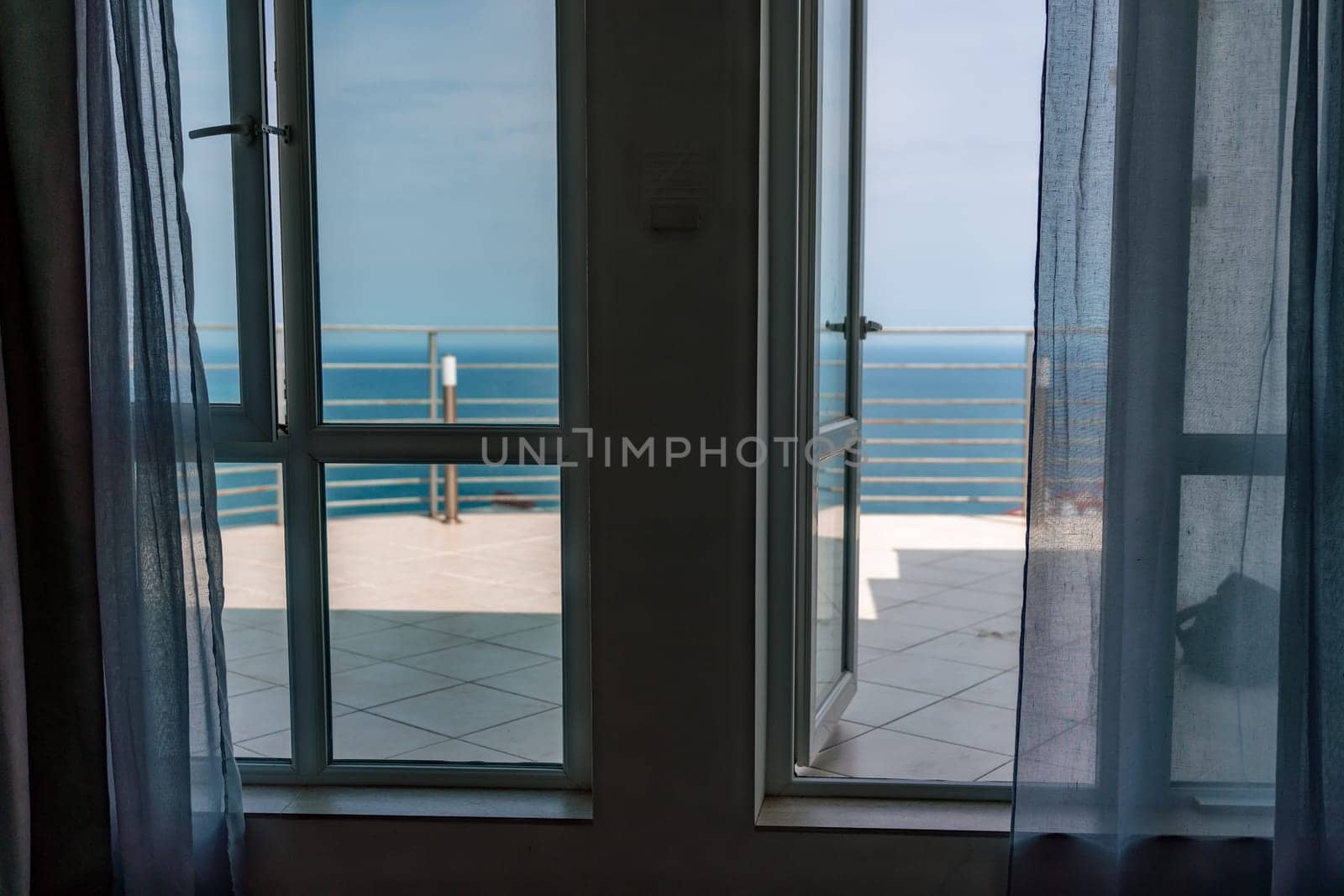 Open window frames a sea view with curtains, leading to a balcony and ocean horizon.