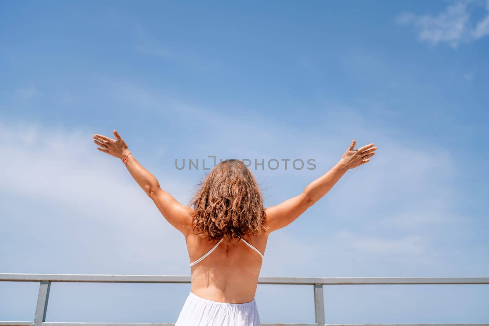 Woman facing away, arms outstretched against a bright blue sky. White dress, curly hair, and distant railing visible. Sunny day.