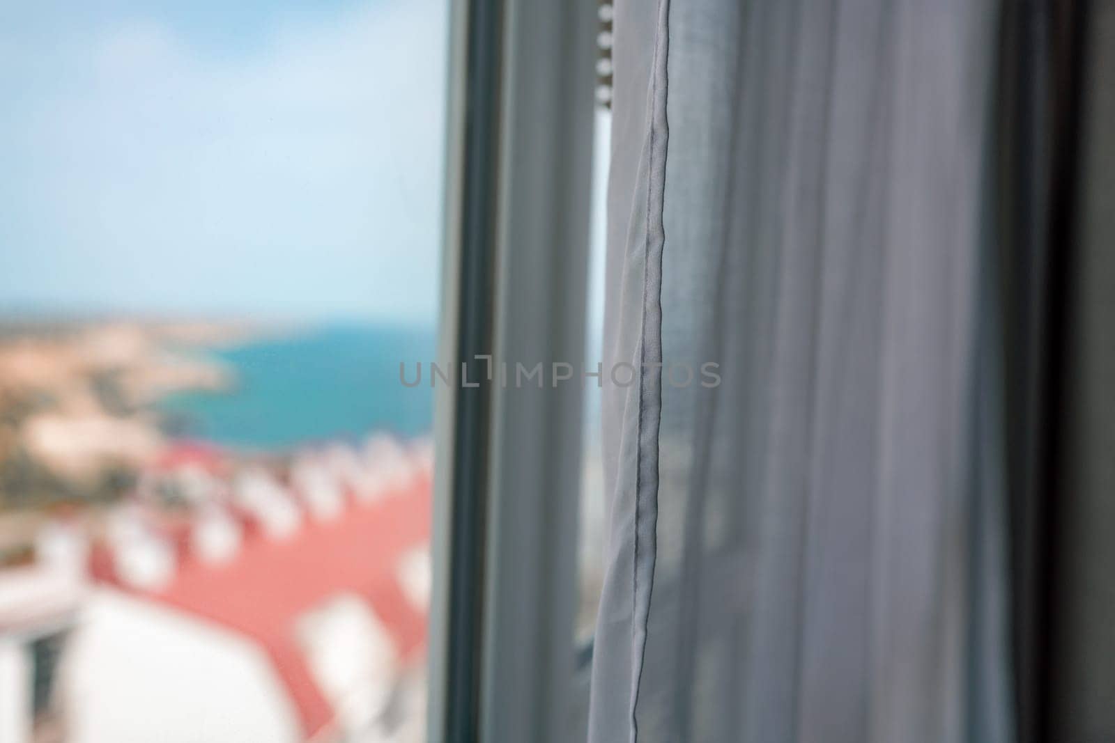 View from a window: A blurred landscape of buildings and blue water seen through a gap in white curtains.