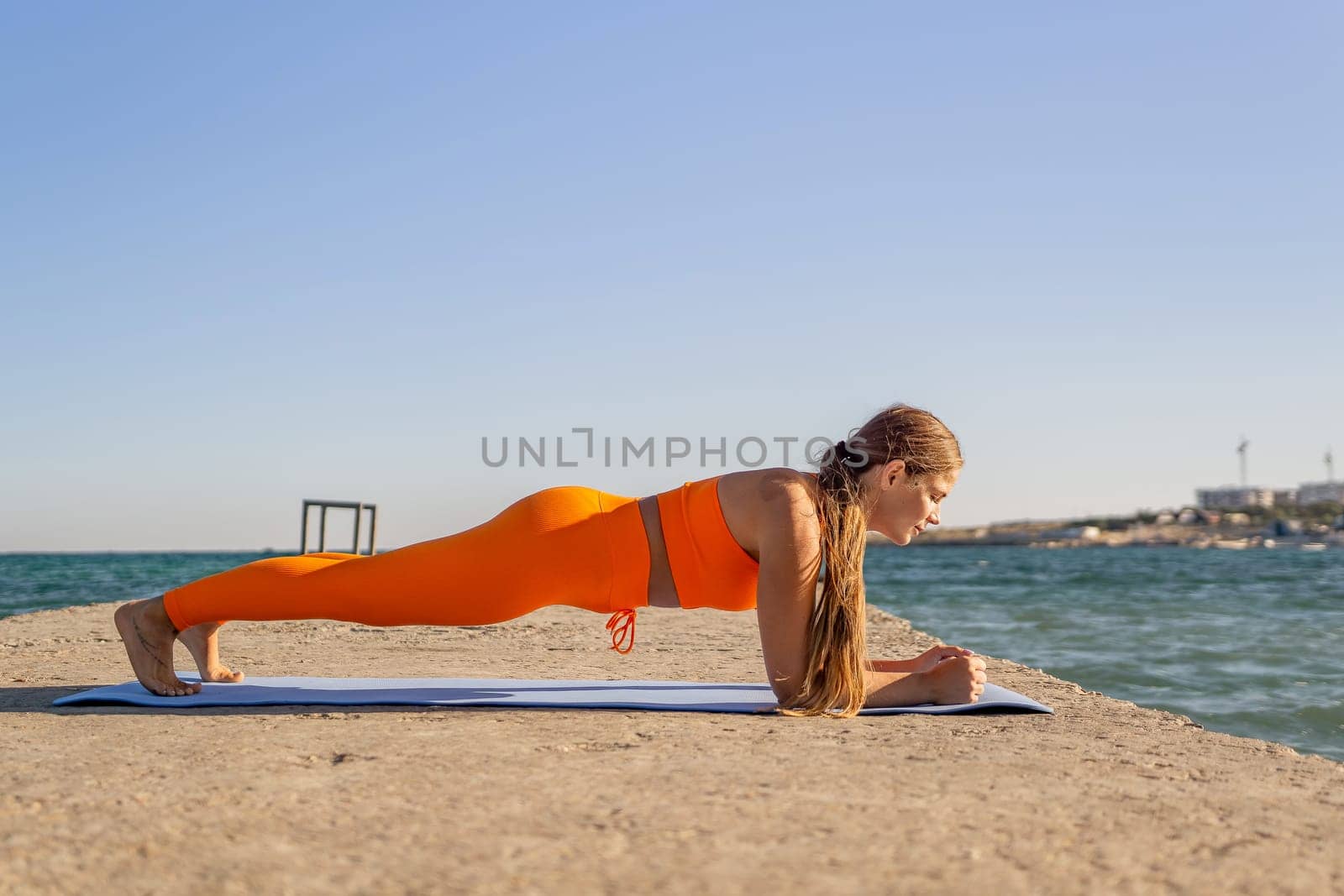 A woman in orange workout clothes performs a plank on a yoga mat on a concrete pier overlooking the ocean.