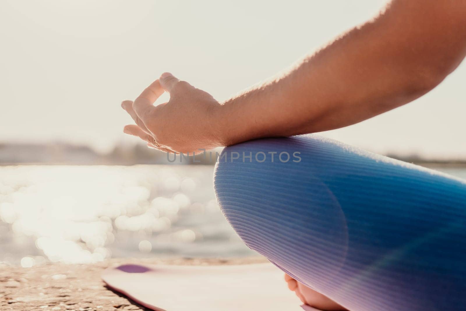 A woman sits in a yoga pose with her legs crossed and hands in a mudra. She is wearing blue leggings and is sitting on a mat. There is a body of water behind her.