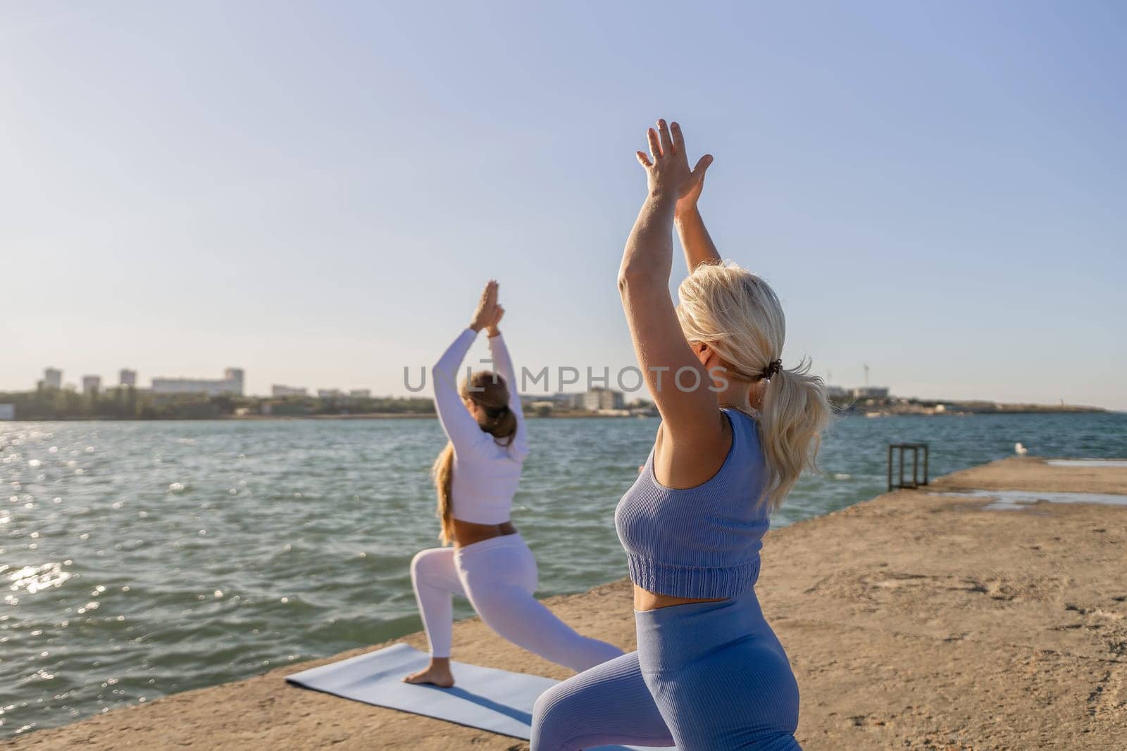 Two people in athletic wear practice yoga on a sandy beach by the water. One is in a lunge with arms raised, the other has their arms raised overhead. The sky is blue and the water is calm.