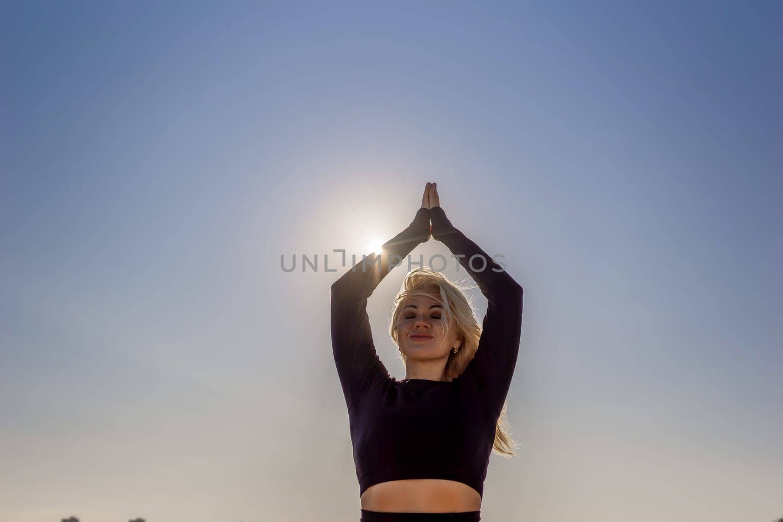 A woman with blonde hair practices yoga outdoors, her hands raised above her head in a prayer position, with the sun shining brightly in the background.
