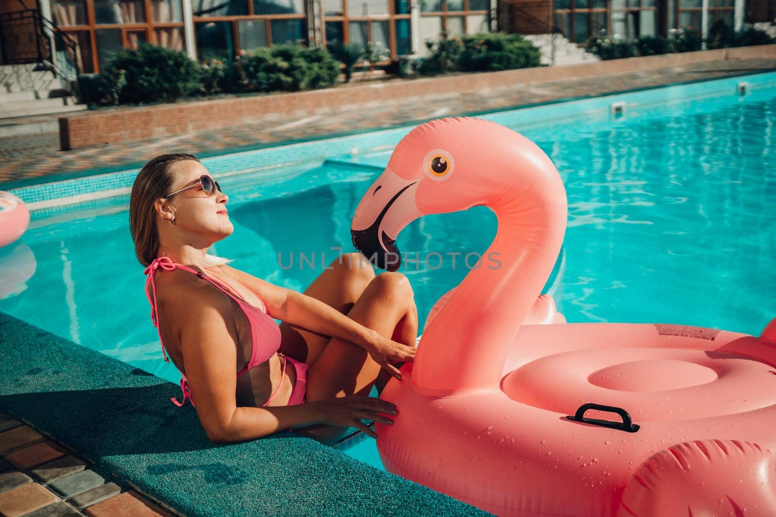 A woman in a pink bikini is sitting on a pink inflatable flamingo in a pool. The scene is playful and fun, with the woman enjoying her time in the water