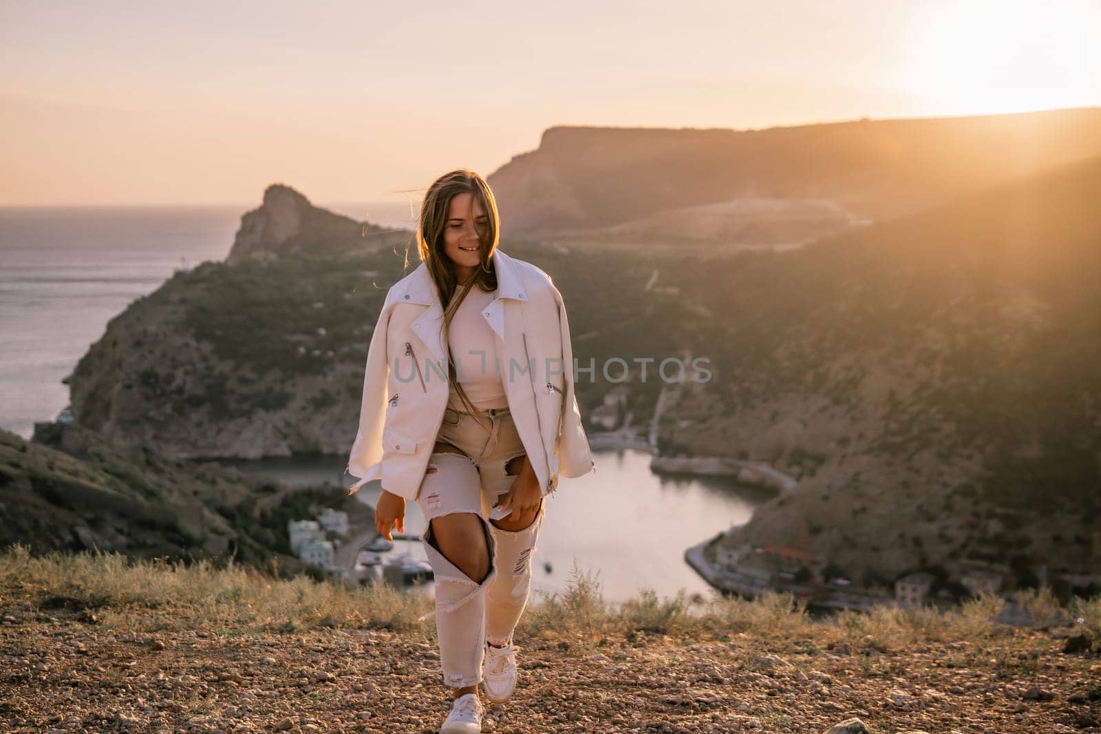 A woman stands on a hill overlooking a body of water. The sun is setting in the background, casting a warm glow over the scene. The woman is enjoying the view and taking in the beauty of the landscape