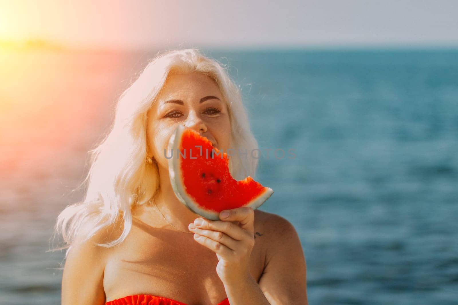Blond woman in red bikini holds a juicy watermelon slice, partially obscuring her face, against a bright blue ocean backdrop. Summer vibes.