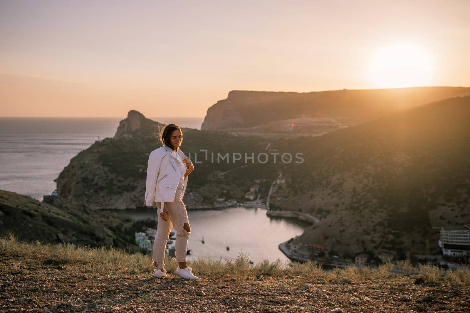 A woman stands on a hill overlooking a body of water. The sun is setting in the background, casting a warm glow over the scene. The woman is enjoying the view and taking in the beauty of the landscape