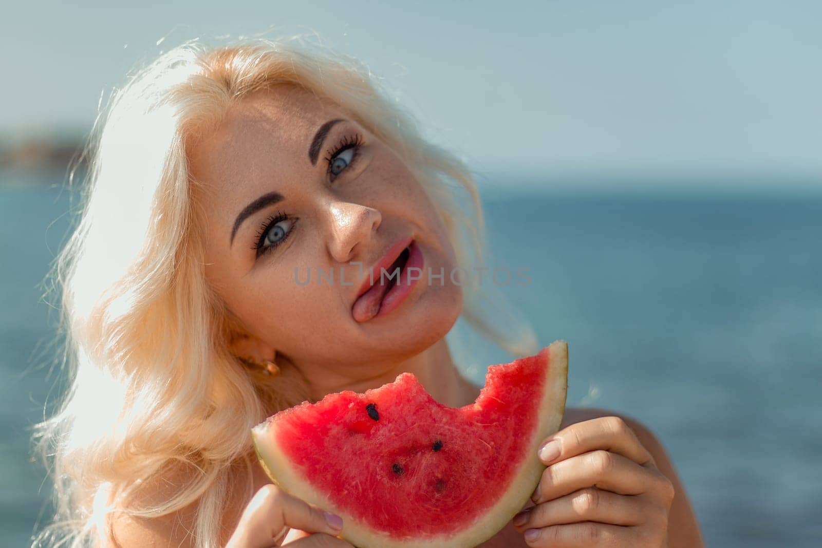 Blonde woman with playful expression, tongue out, eating a slice of watermelon near the ocean. Summer vibes.