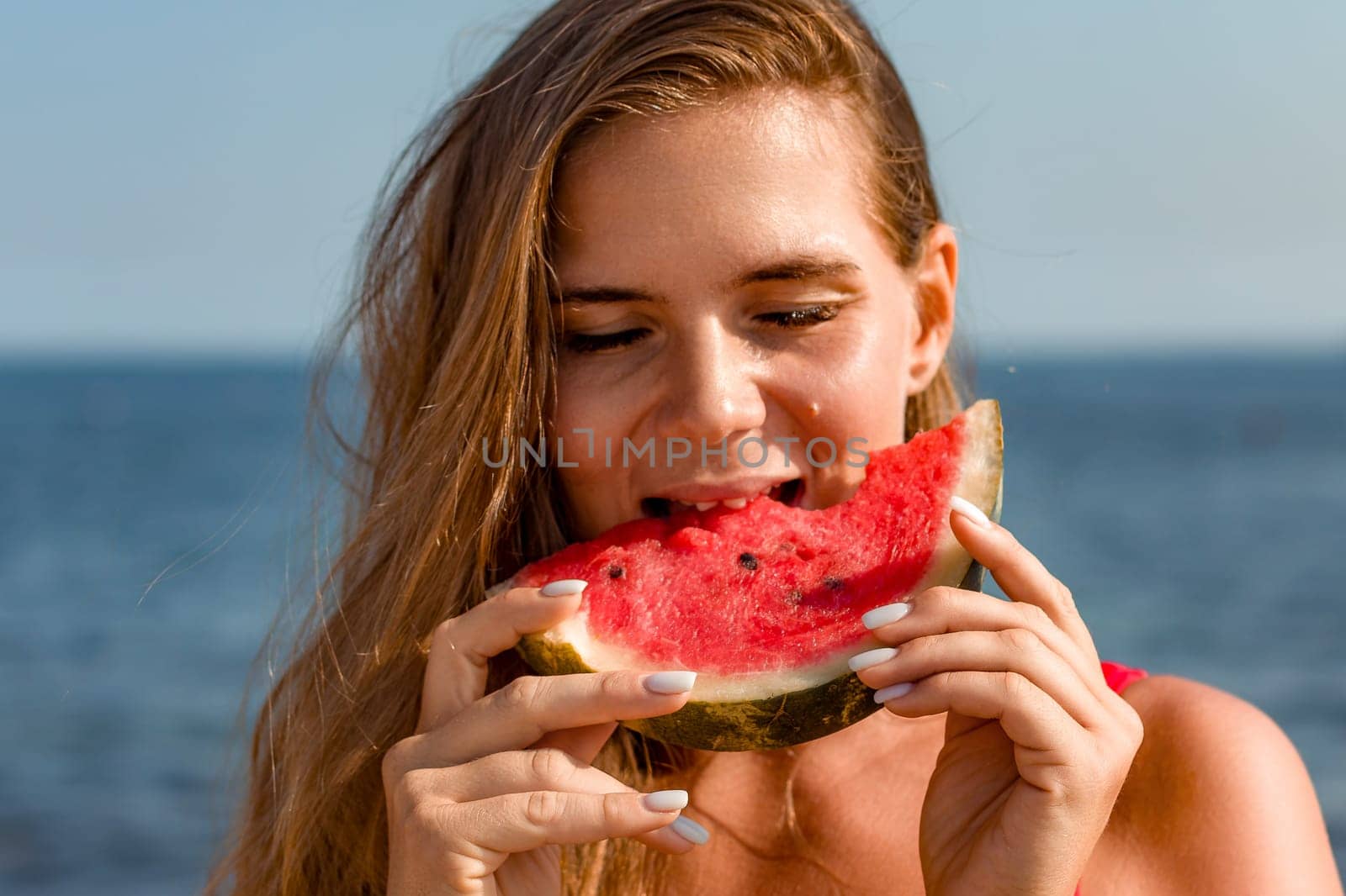 Woman with long brown hair enjoys a juicy slice of watermelon at the beach. Sunlight illuminates her face as she bites into the refreshing fruit.