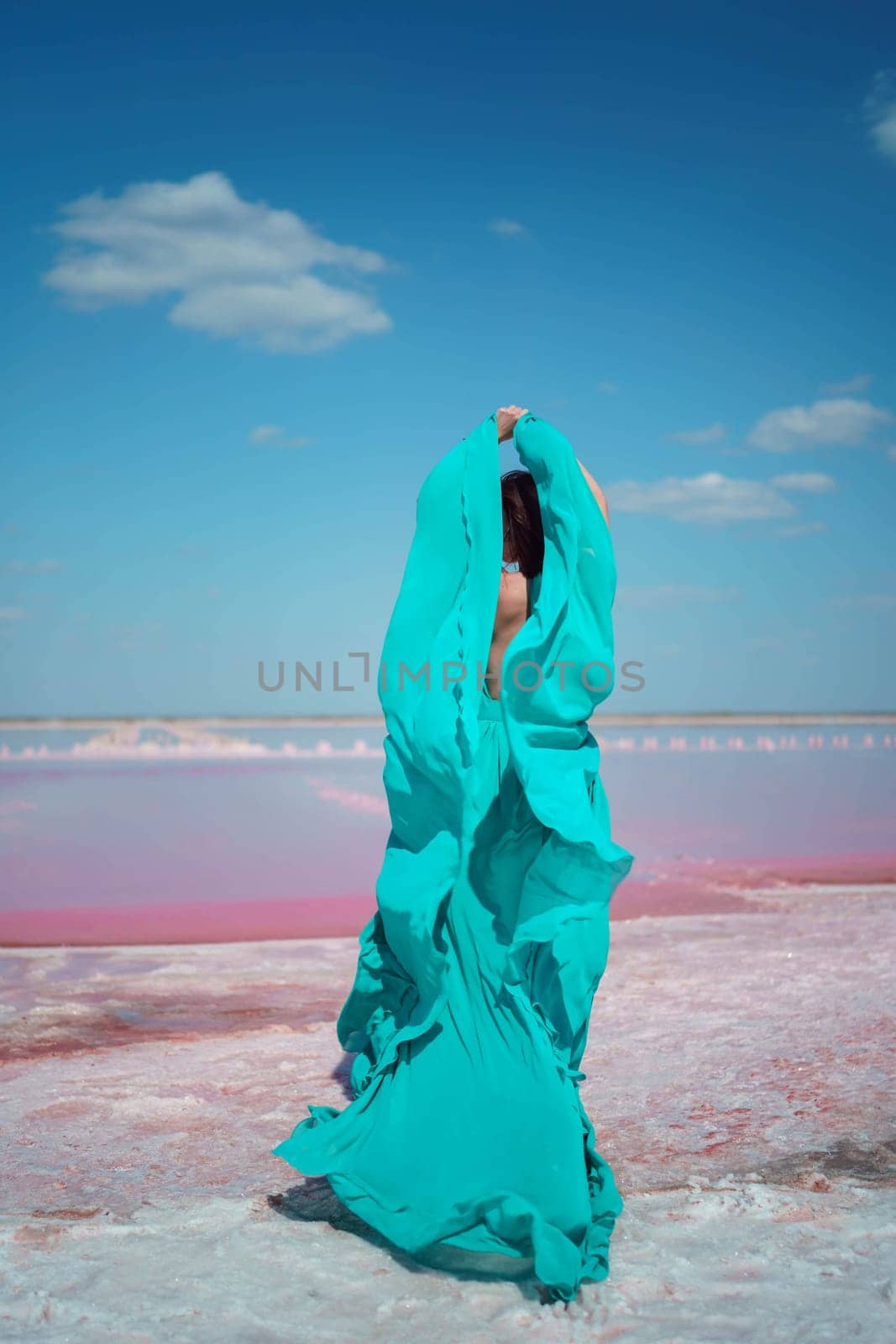 Woman in teal fabric, arms raised, stands on pink salt lake shore under a vibrant blue sky. Windblown fabric billows dramatically.
