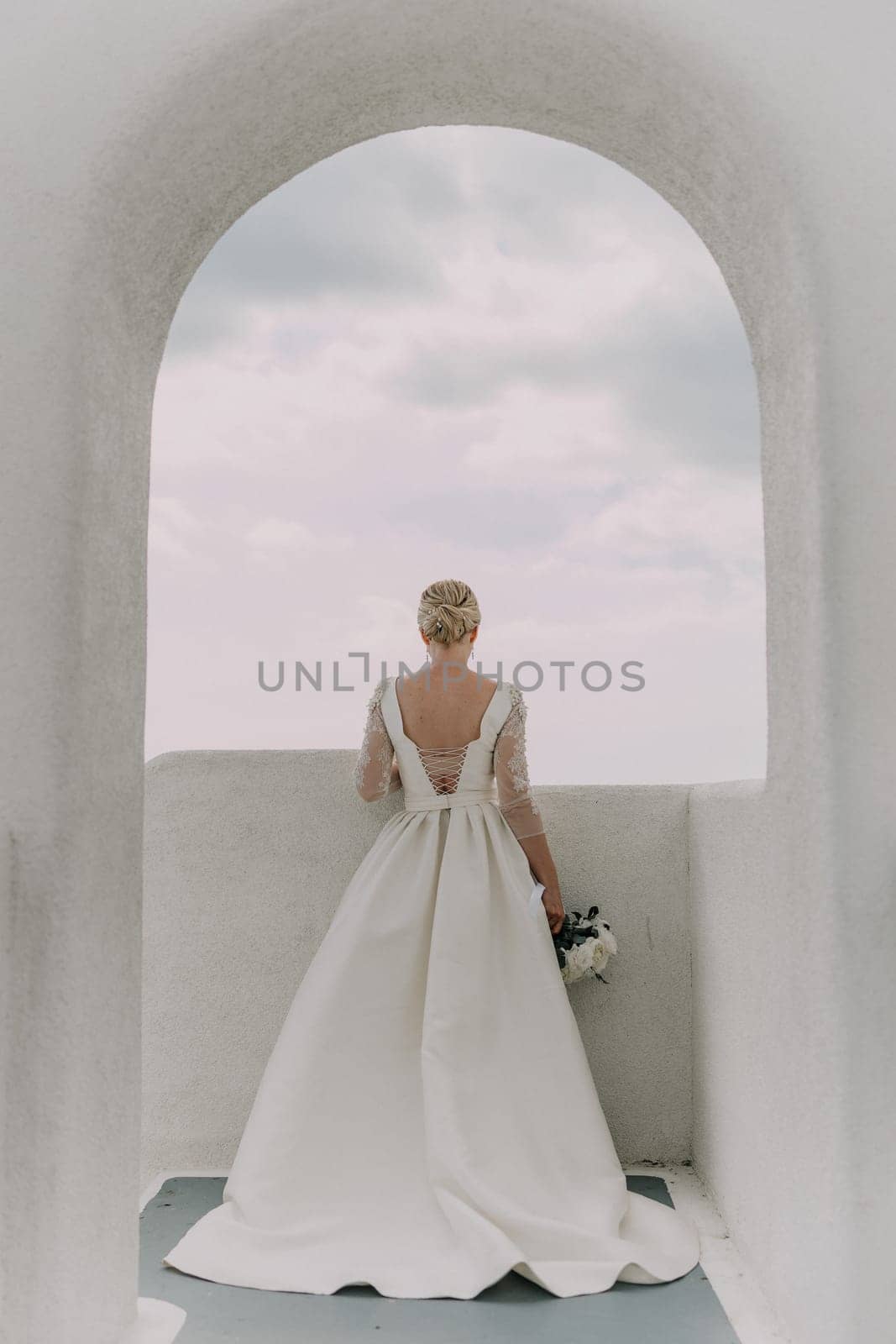 A bride stands in front of an archway, holding a bouquet of flowers. The archway is white and the sky is cloudy. The bride is wearing a white dress and her hair is styled in a bun