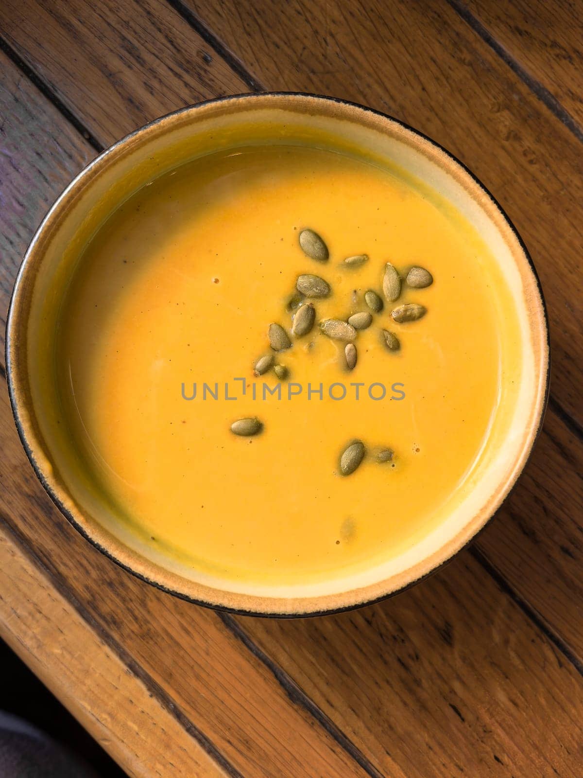 Creamy pumpkin soup served in a bowl, garnished with pumpkin seeds on top. The soup is placed on a wooden table. Photo taken with a smartphone.