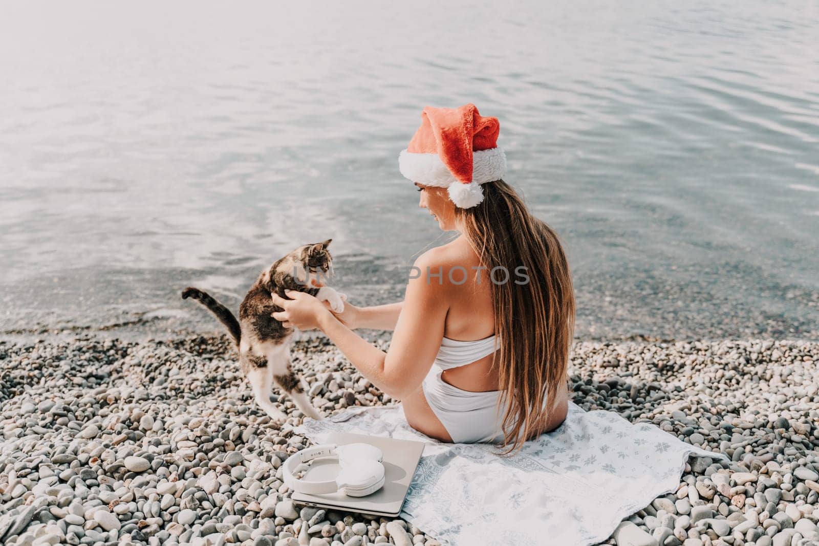 A woman wearing a red Santa hat sits on a beach towel. She is wearing a bikini and has a laptop on her lap