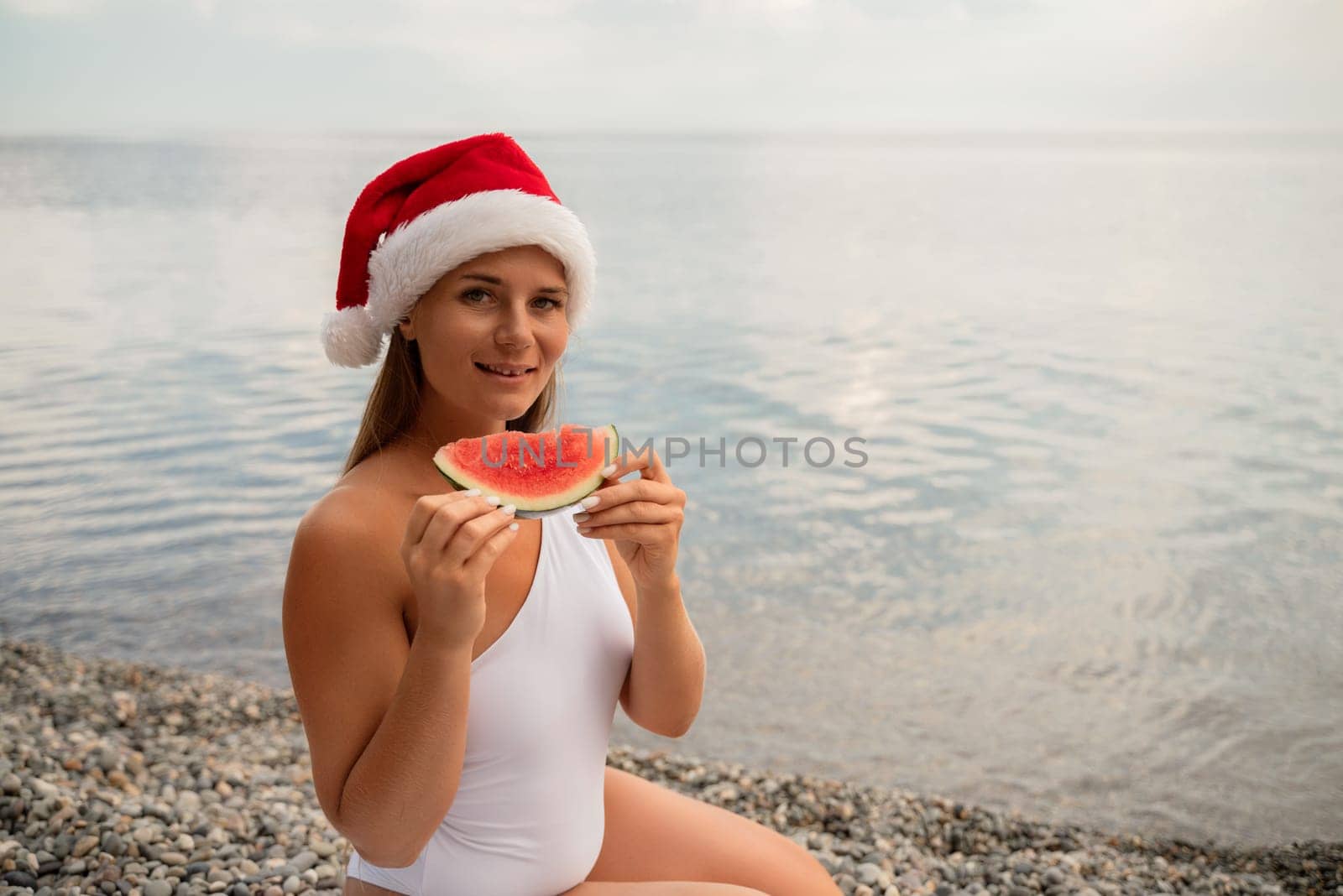 A woman is eating a watermelon while wearing a red santa hat. Scene is lighthearted and festive, as the woman is enjoying a refreshing snack on a sunny day