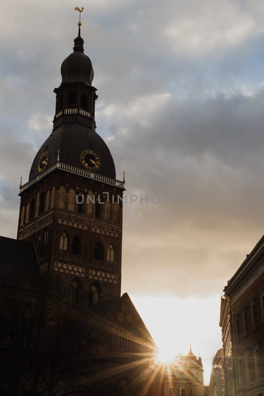 Historic Church Tower with Sunset in the Background.A striking view of a historic church tower silhouetted against a dramatic sky at sunset. The golden sunlight peeks through surrounding buildings, casting a warm glow