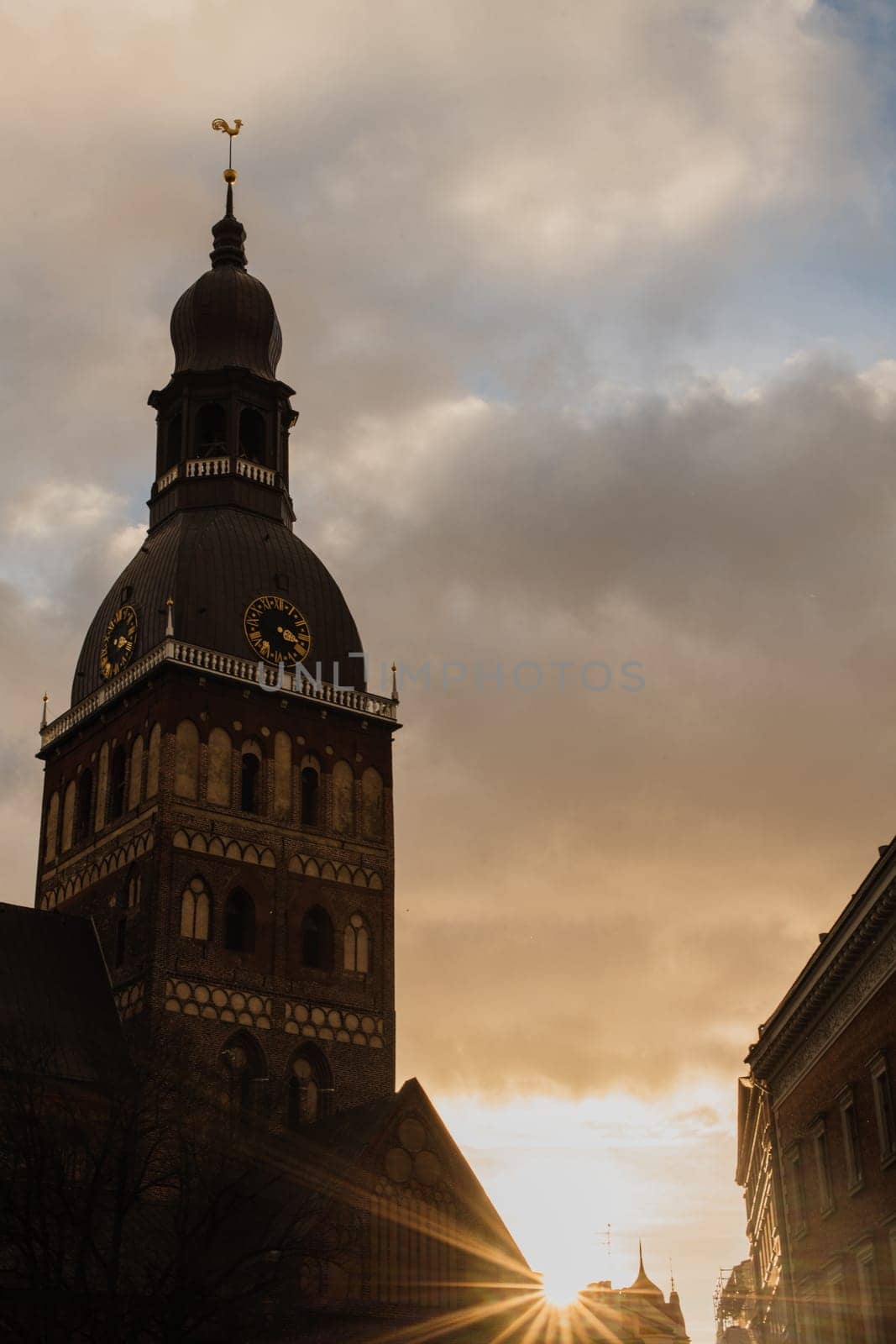 Historic Church Tower with Sunset in the Background.A striking view of a historic church tower silhouetted against a dramatic sky at sunset. The golden sunlight peeks through surrounding buildings, casting a warm glow