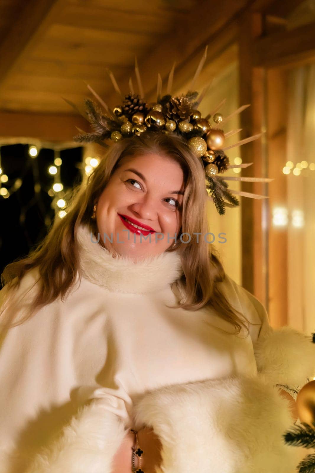 A woman with a festive crown of gold ornaments and branches smiles at the camera. She wears a white fur coat and red lipstick. The background is blurry, but appears to be a warm, decorated space.