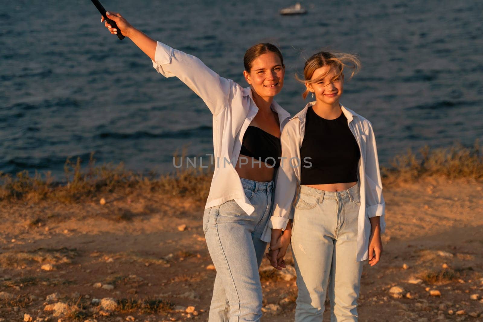 Two young women in white shirts and jeans stand near a body of water, one holding a selfie stick. They are smiling and looking at the camera.