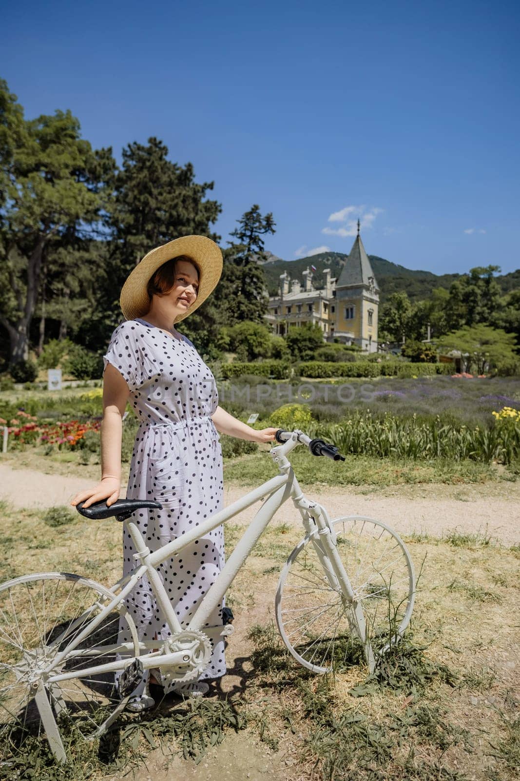 A woman is standing next to a white bicycle. She is wearing a white dress and a straw hat. The scene is set in a park with a house in the background. The woman is enjoying her time outdoors