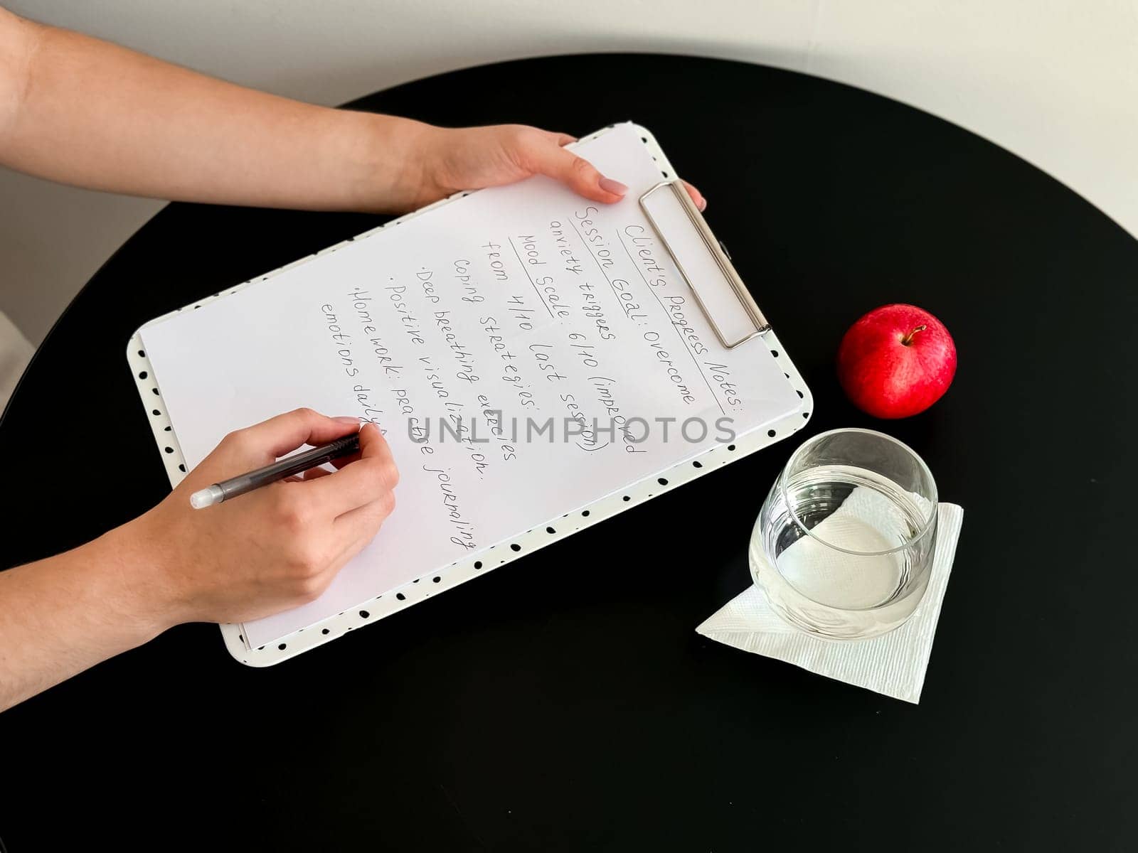 Female writing on clipboard with notes next to apple and glass on round black table.